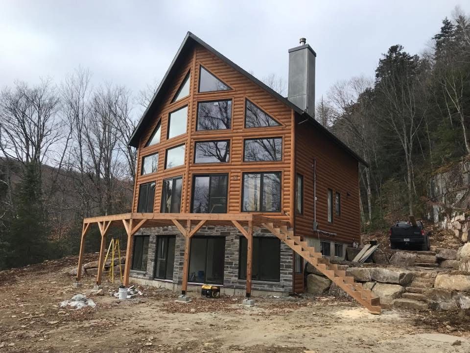Un chalet en rondins de deux étages, avec de grandes fenêtres et une terrasse en bois, se dresse contre un flanc de montagne boisé et rocailleux, sous un ciel gris.