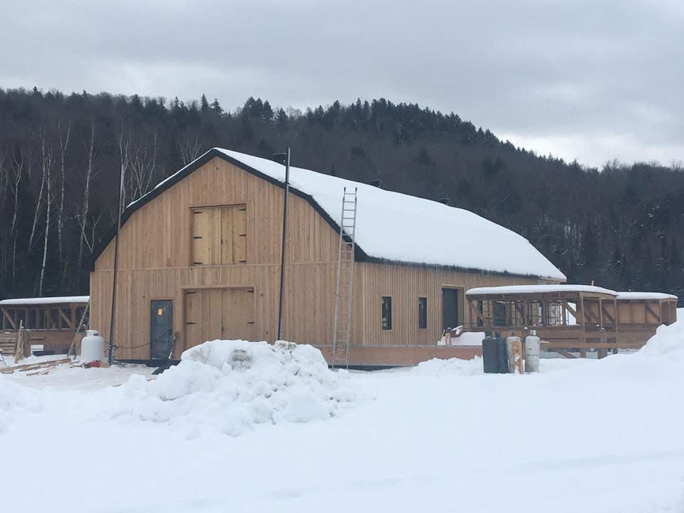 Une grande grange en bois clair, au toit enneigé, se dresse dans un paysage rural enneigé, adossée à une sombre ligne d'arbres.