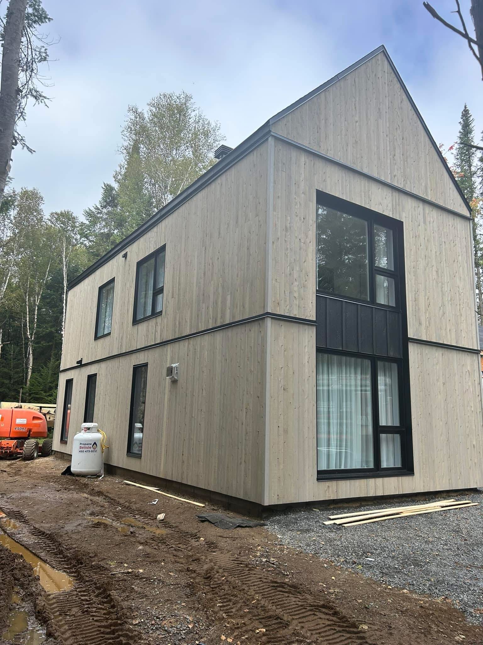 Une maison à deux étages avec un bardage en bois clair vertical et des cadres de fenêtres noirs, située sur un terrain boueux en chantier.