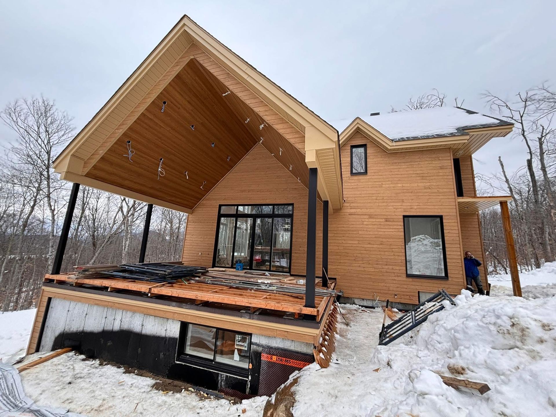 Une maison à deux étages en construction, avec un bardage en bois clair, une grande terrasse couverte et un jardin boisé enneigé.
