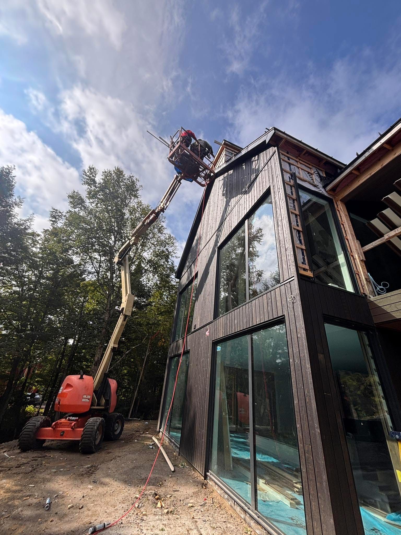 Un ouvrier du bâtiment, à bord d'une nacelle élévatrice, atteint le sommet d'une maison moderne en construction, aux façades sombres et aux parois de verre.