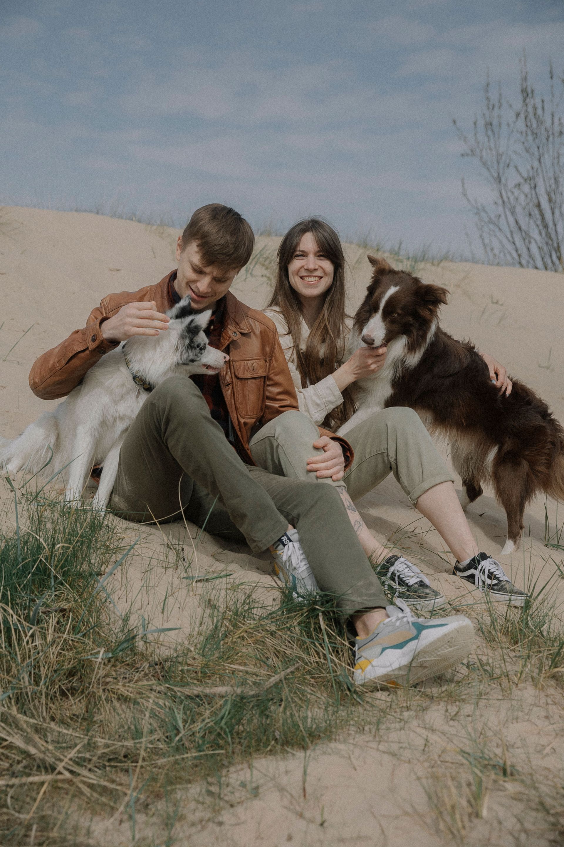 Twee mensen zitten met twee honden in de duinen en glimlachen buiten onder een bewolkte hemel.