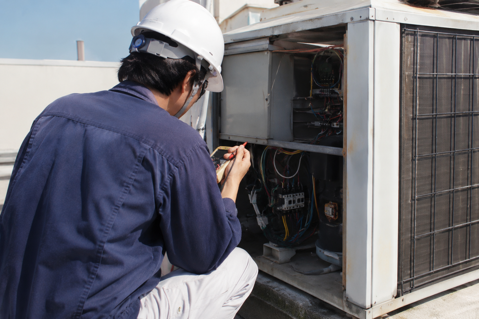 Technician in hard hat inspecting an open HVAC unit on a rooftop.