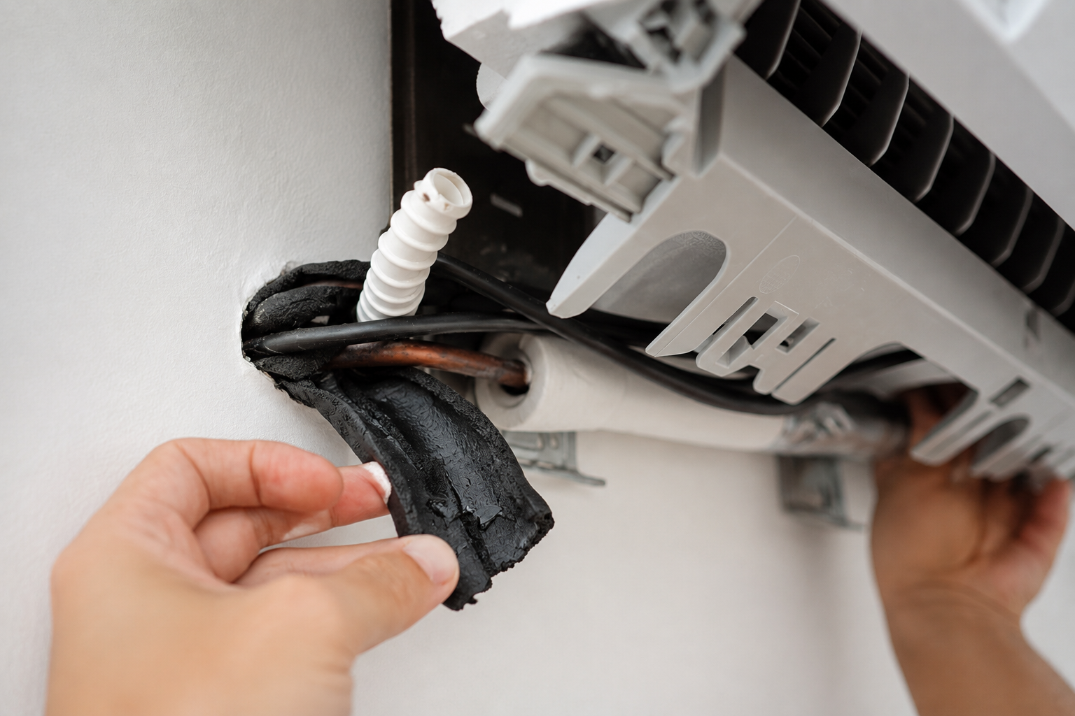 Hands installing an air conditioner unit with exposed wires and tubing on a wall