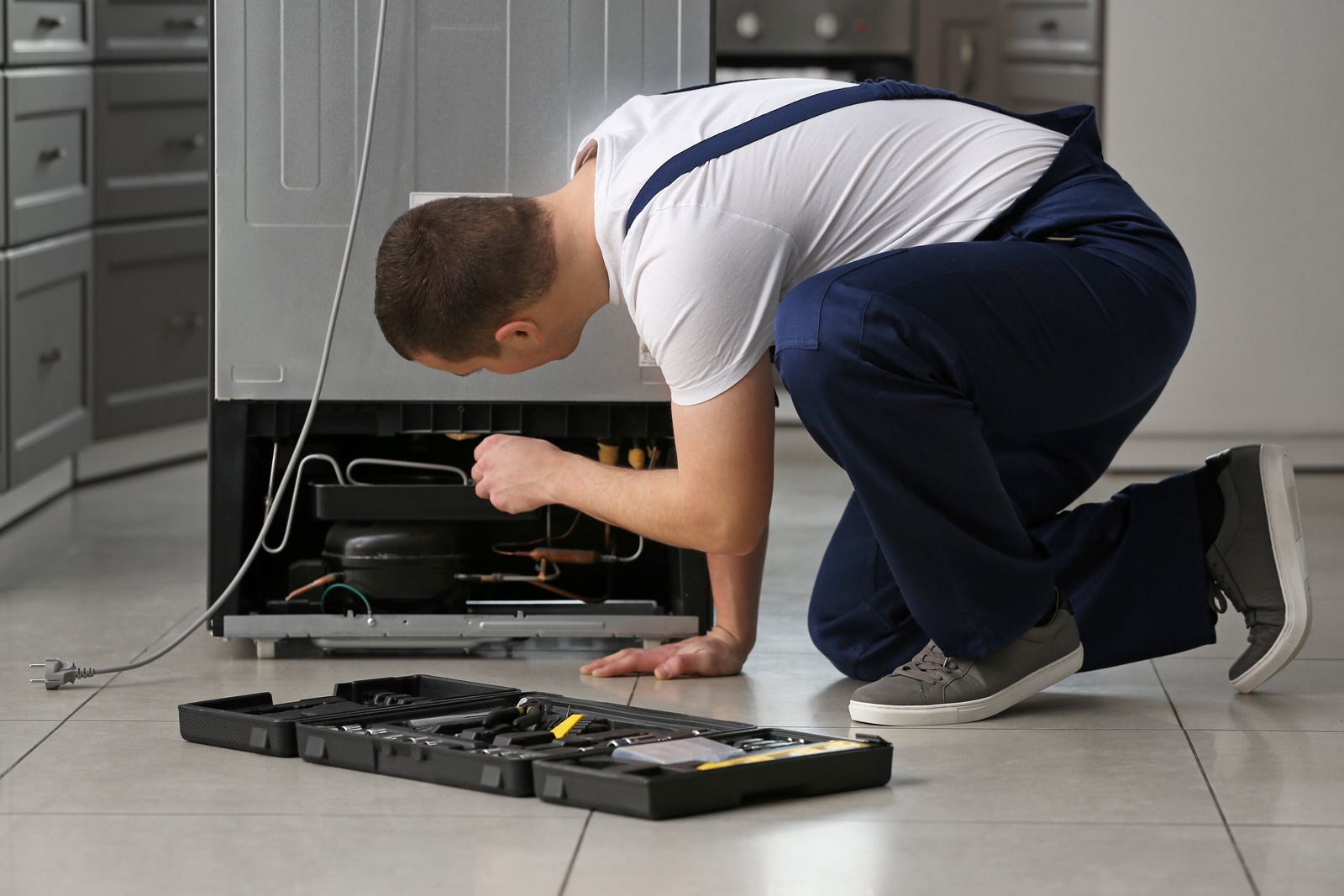 Person crouching to repair an open dishwasher with tools spread on the kitchen floor.