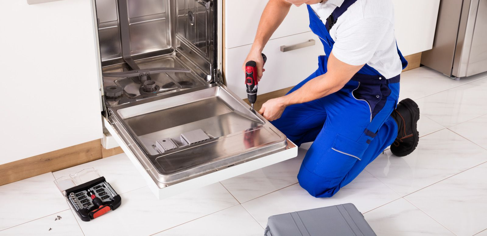 Technician kneels by an open dishwasher, using a screwdriver to repair the lower panel.