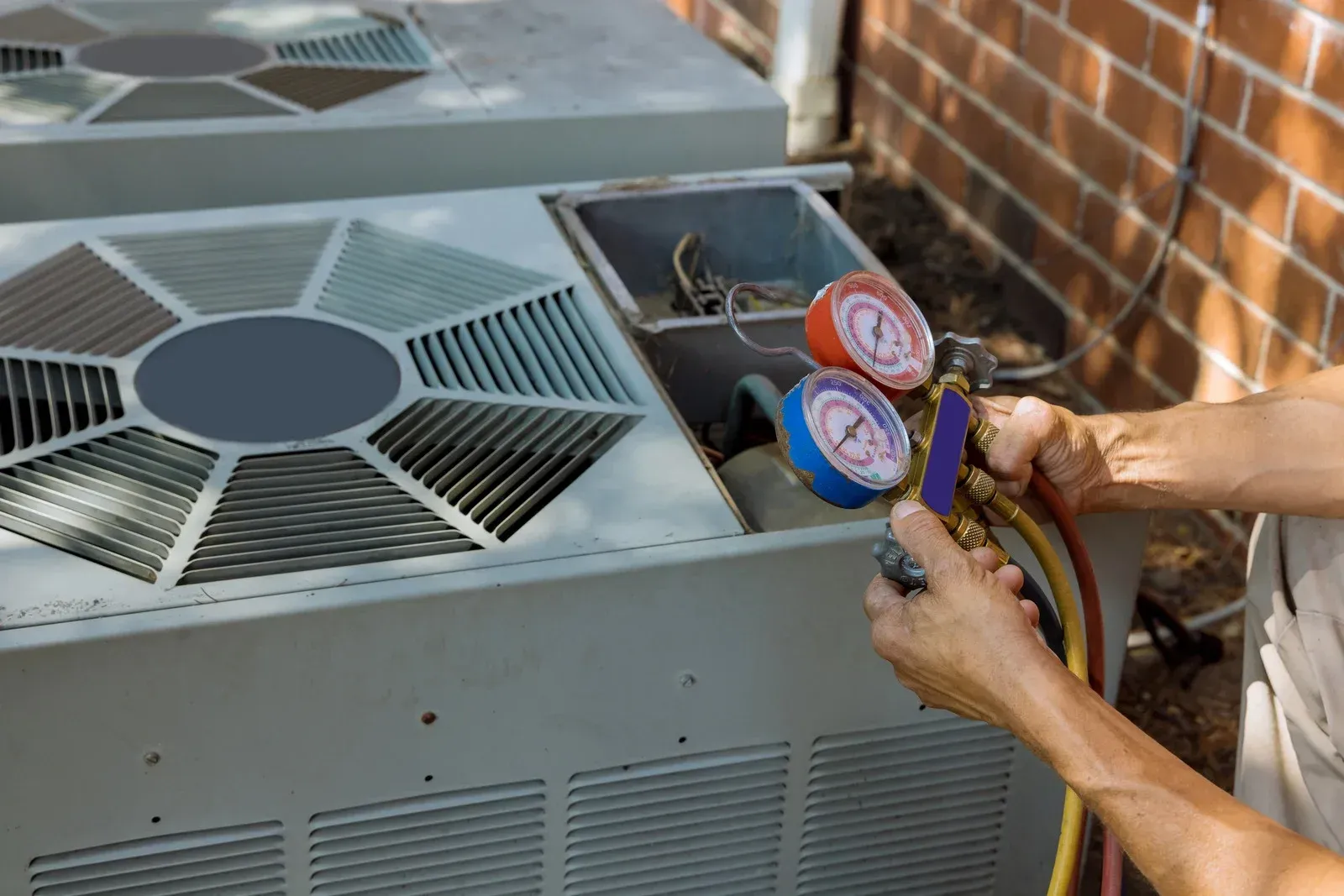 Technician checking HVAC refrigerant gauges on a rooftop unit beside a brick wall