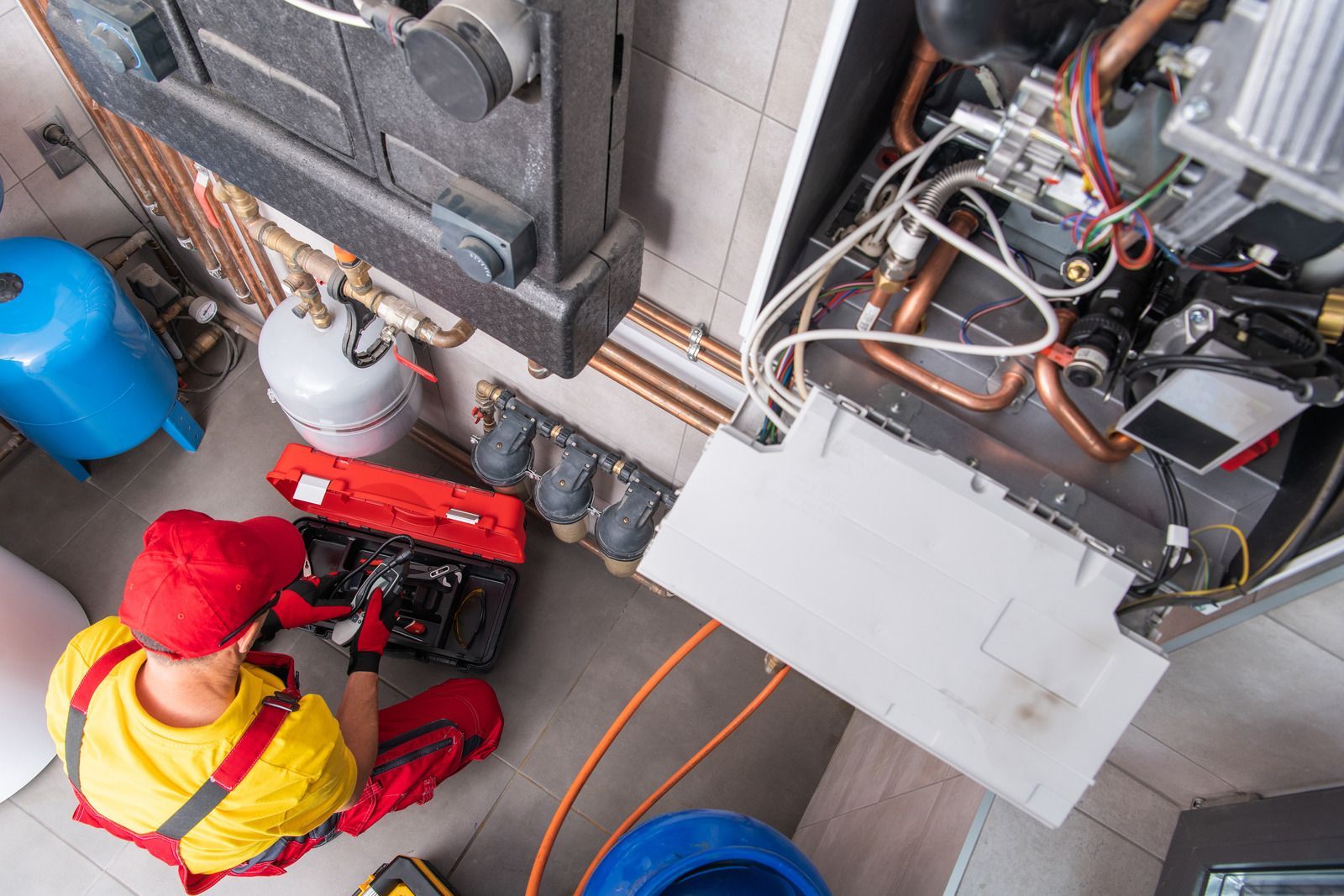 Technician in yellow shirt and red hat inspecting HVAC unit with tools in a utility room