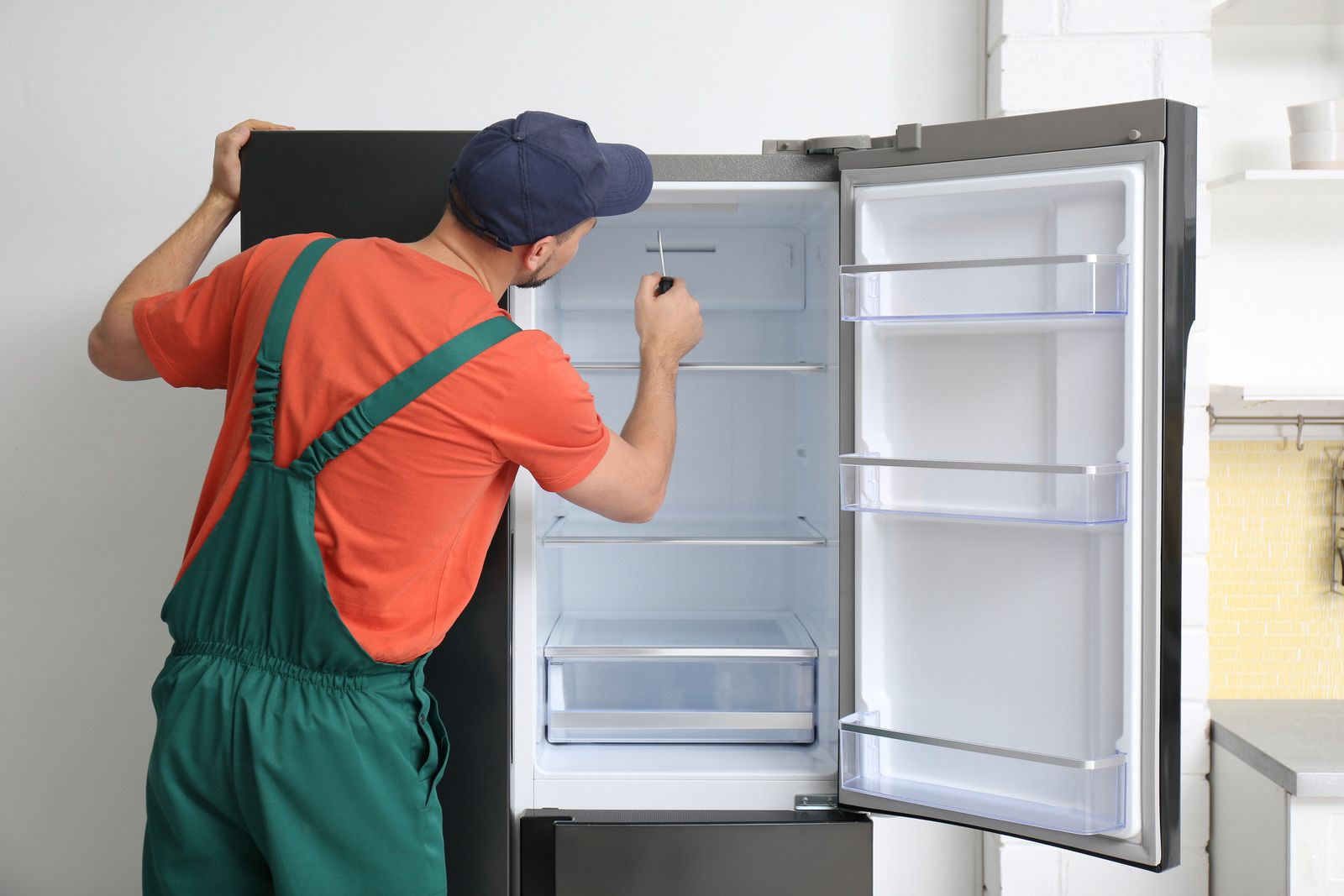 Technician in green overalls installing a refrigerator in a kitchen, door open and shelves visible