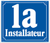Blaues Schild mit weißer Aufschrift „1a“ und „Installateur“, umrahmt von einem dekorativen Rand.