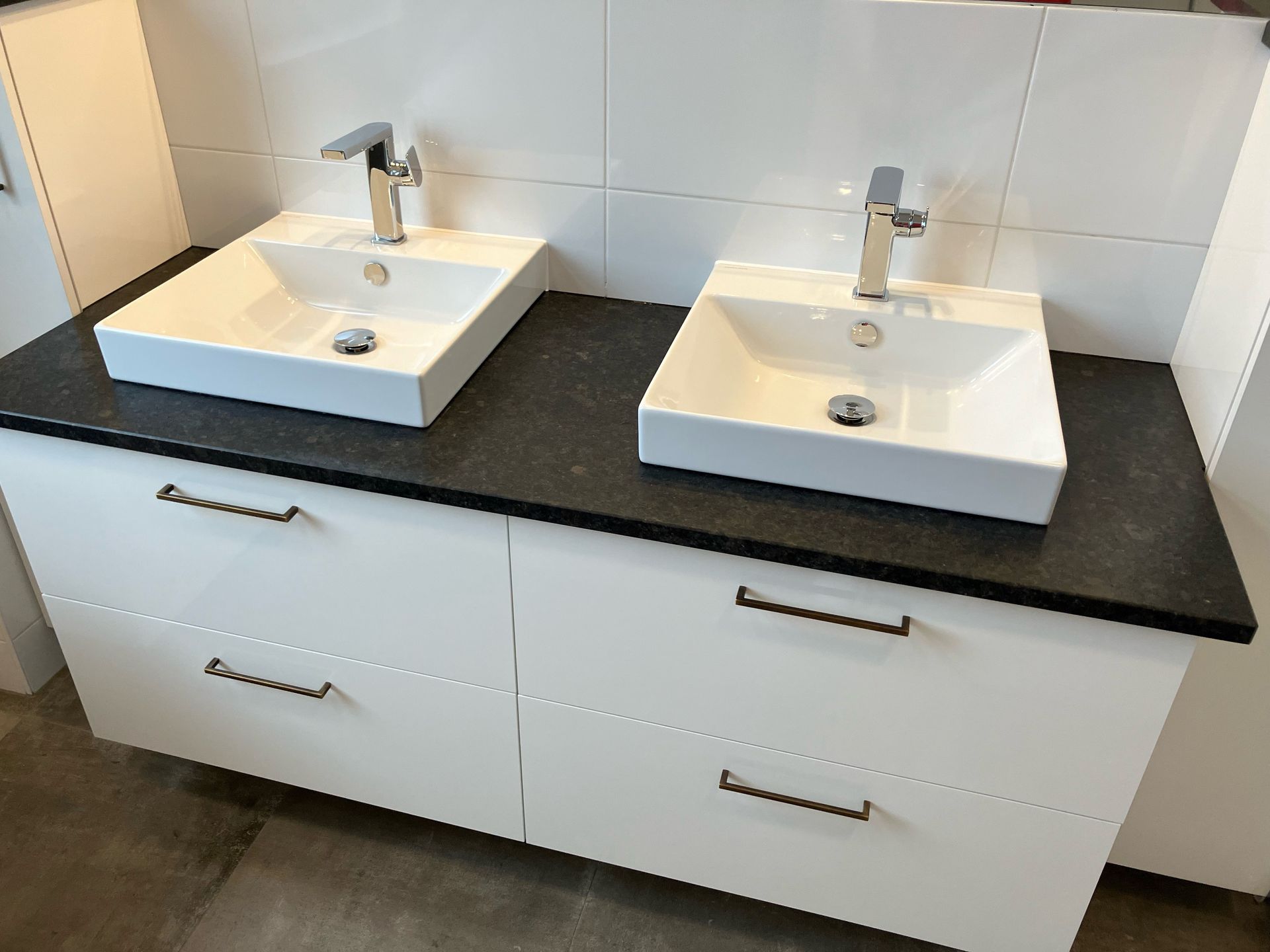 Badezimmerschrank mit zwei weißen Waschbecken, schwarzer Arbeitsplatte, weißen Schränken und verchromten Armaturen.