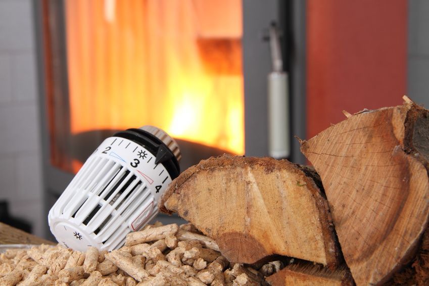 Thermostat neben Holzpellets und Brennholz, im Hintergrund ein brennender Kamin.