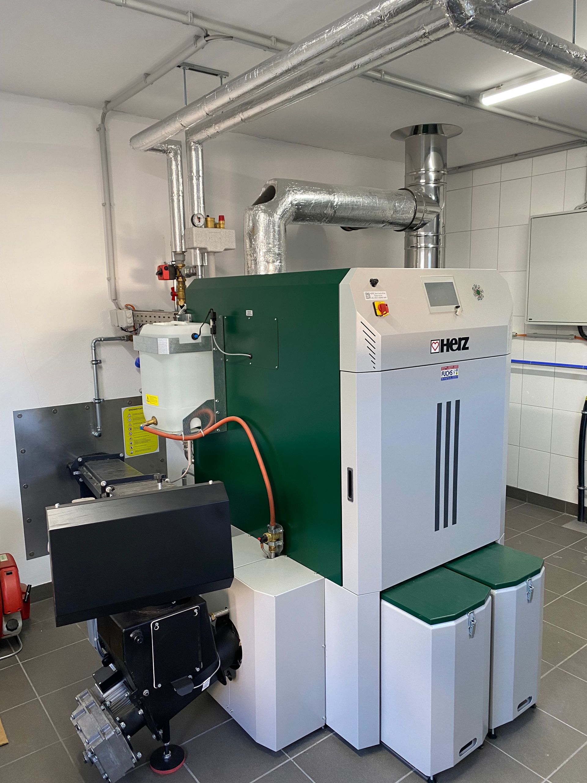 Industrielle Heizanlage mit grünen und weißen Bauteilen, Rohrleitungen und zwei grünen Behältern in einem Technikraum.