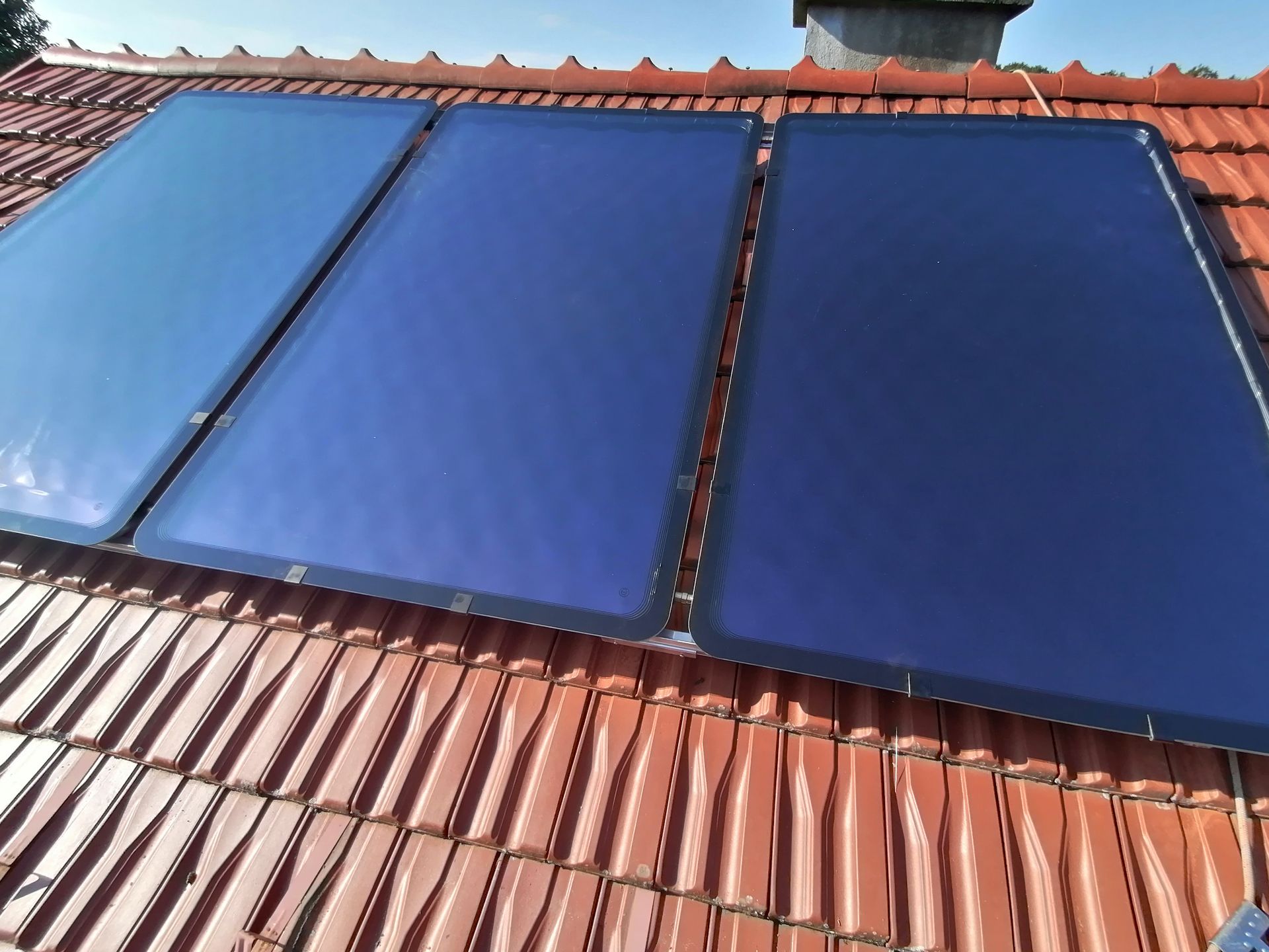 Drei blaue Solarpaneele auf einem roten Ziegeldach vor klarem, blauem Himmel.