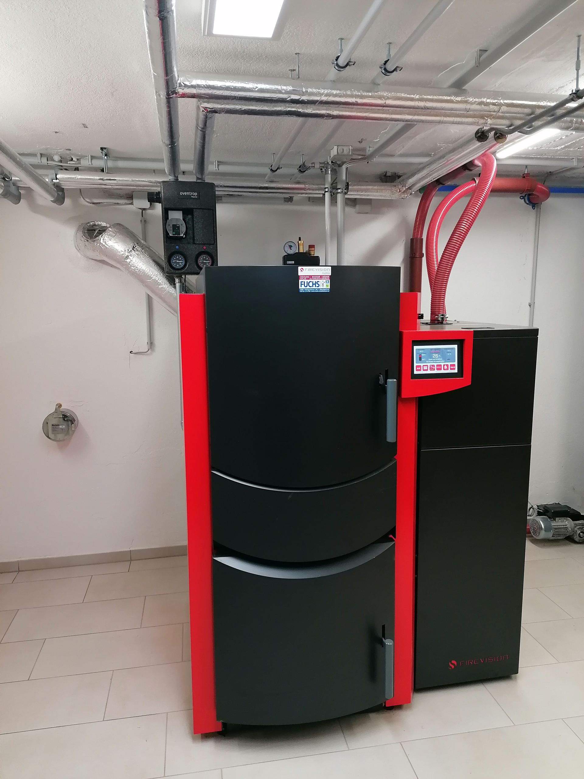 Schwarzer und roter Heizkessel in einem Hauswirtschaftsraum mit Rohren und Digitalanzeige.
