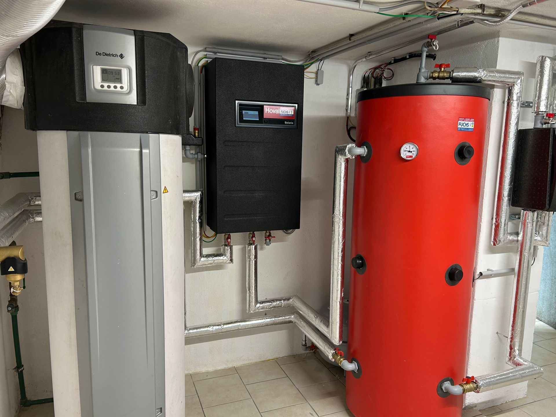 Komponenten des Heizsystems: ein grauer Wassererhitzer, ein schwarzer Schaltkasten und ein roter Wassertank mit Rohren.