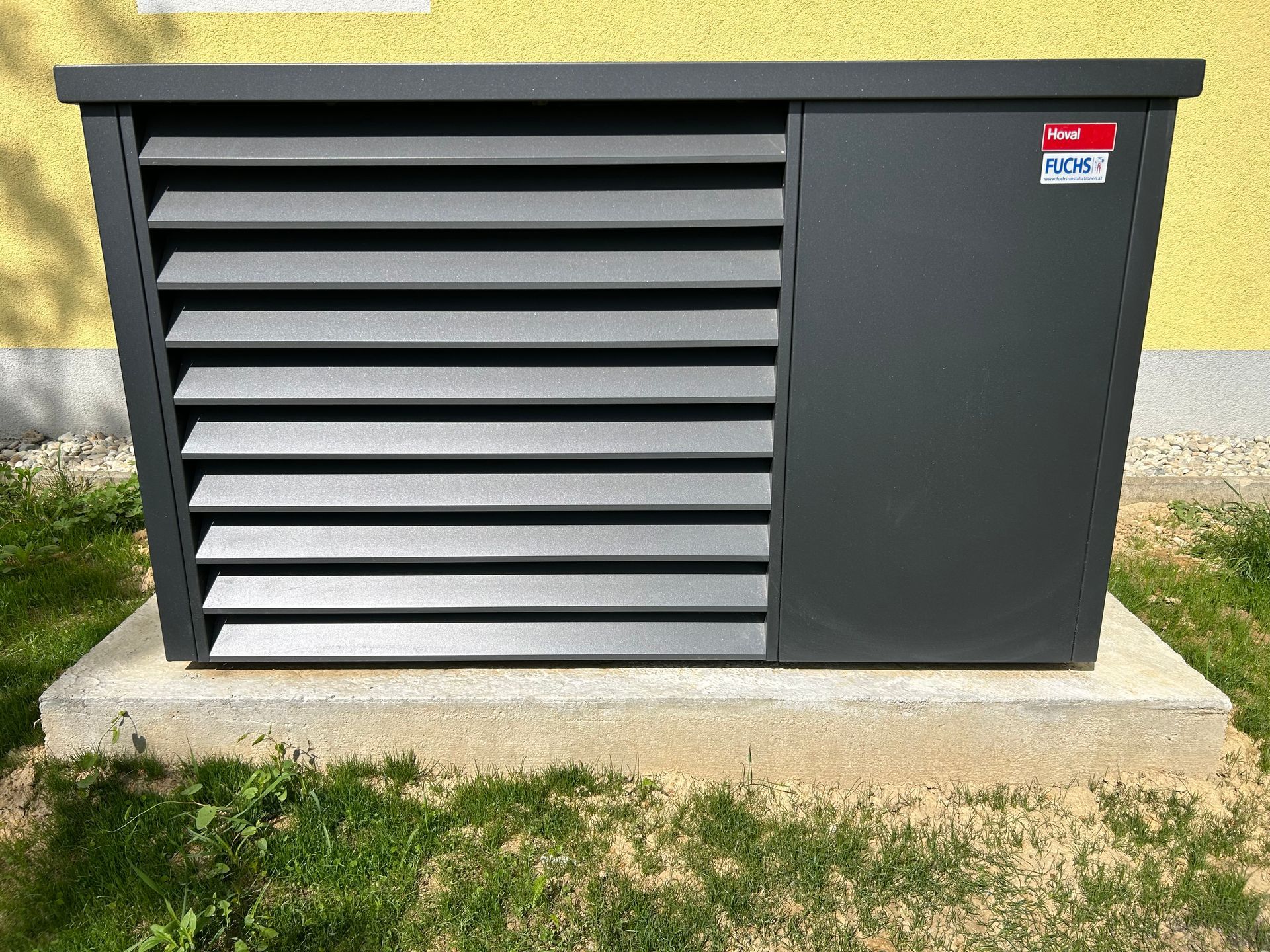 Dunkelgraue Wärmepumpe mit horizontalen Lamellen, montiert auf einem Betonsockel, neben einer gelben Wand.
