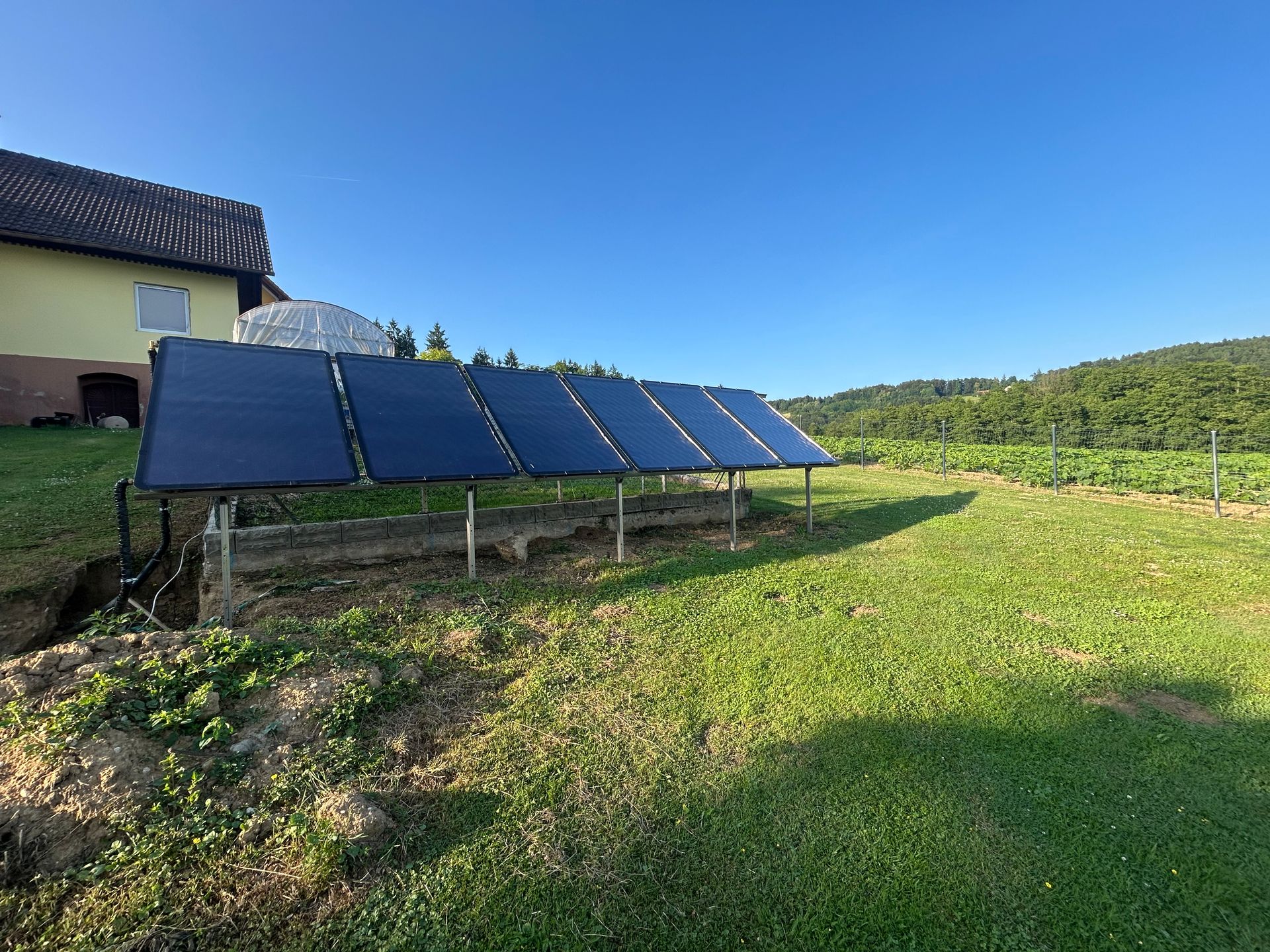 Solarpaneele in einem grasbewachsenen Hof, im Hintergrund ein Haus und Bäume unter blauem Himmel.