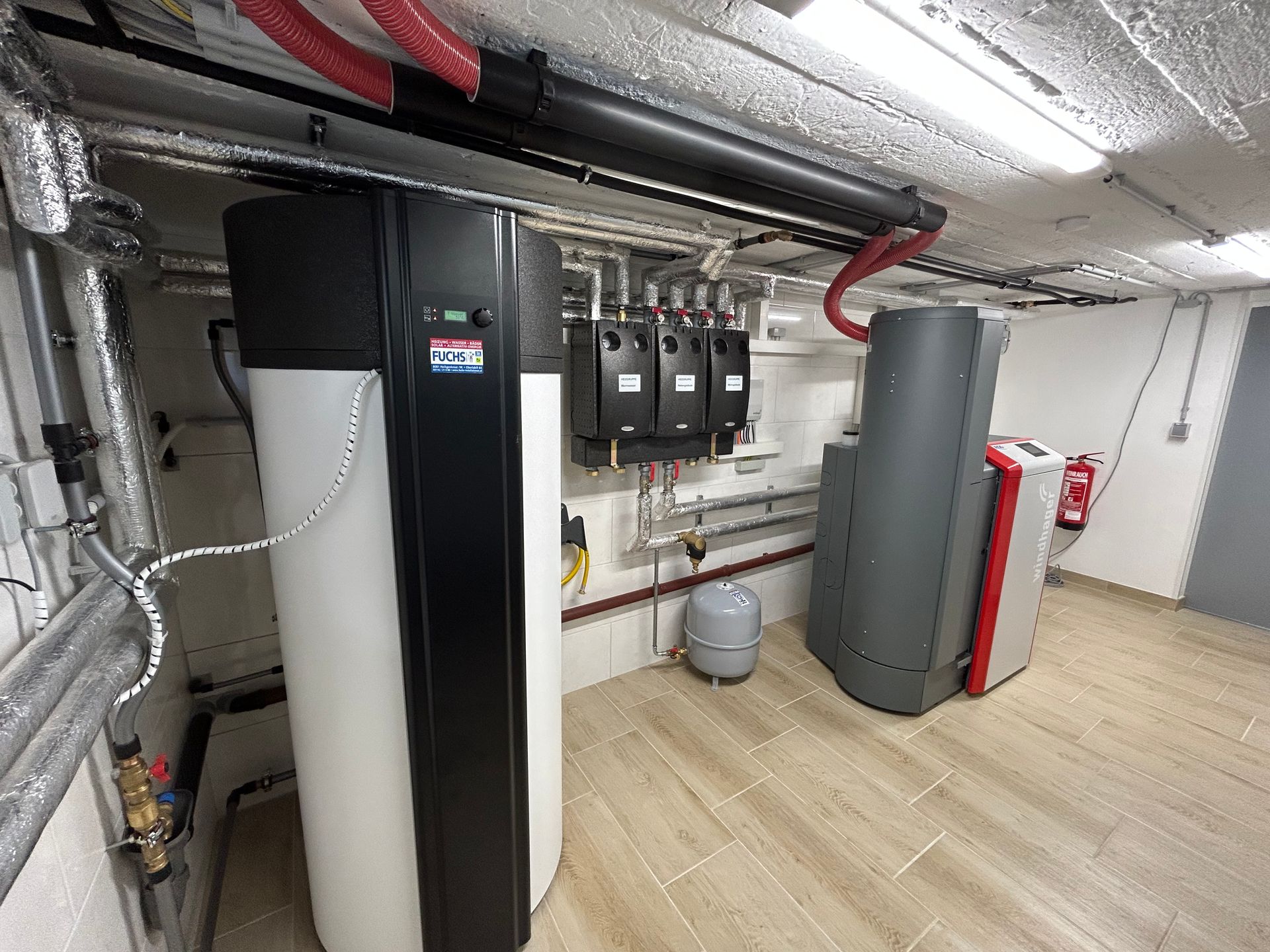 Ein Technikraum im Kellergeschoss mit Heizungs- und Sanitäranlagen; Rohre, Tanks und ein Feuerlöscher sind sichtbar.