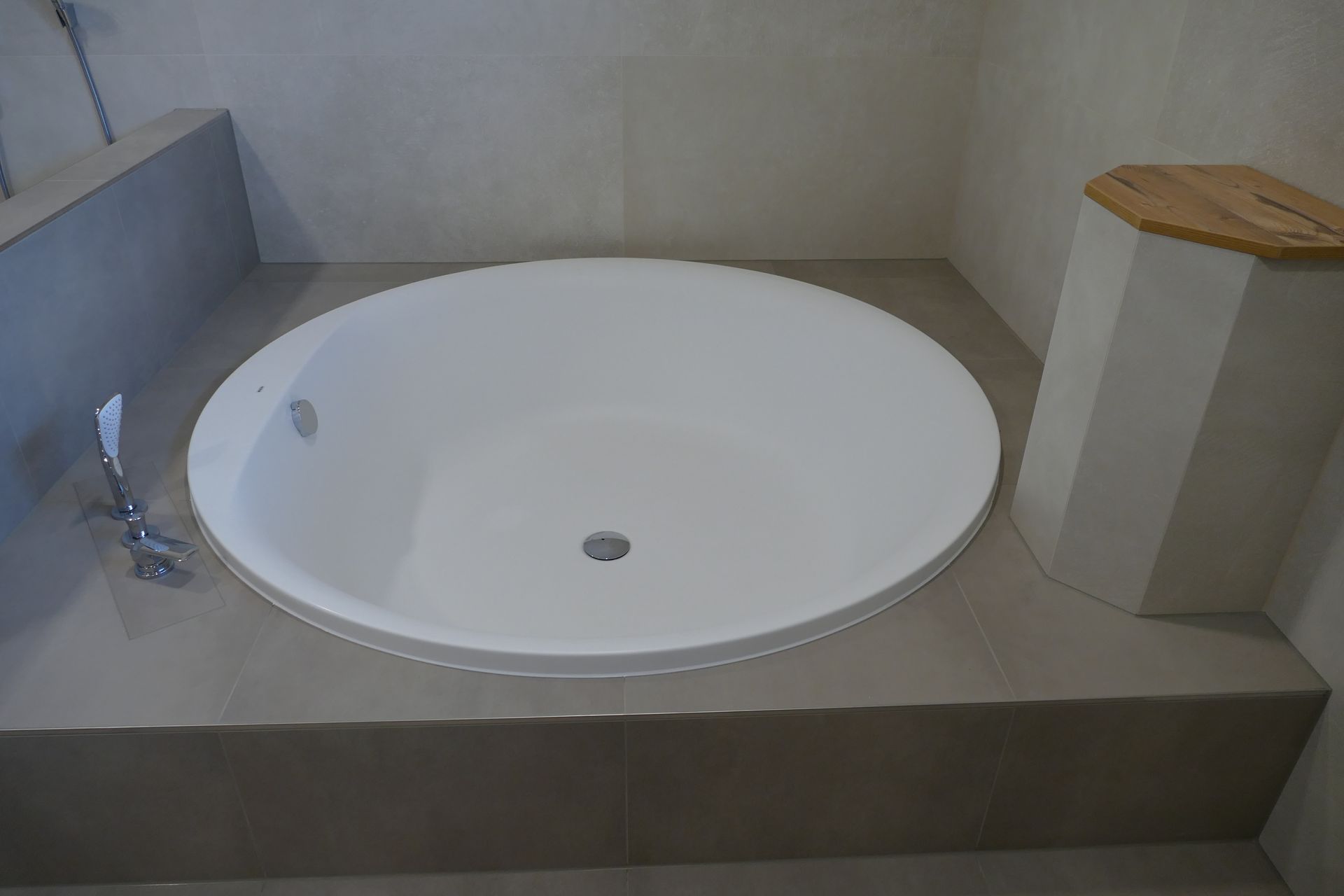 Weiße, runde Badewanne, eingelassen in ein hellgraues Fliesenpodest, daneben ein dekorativer Holzschemel.