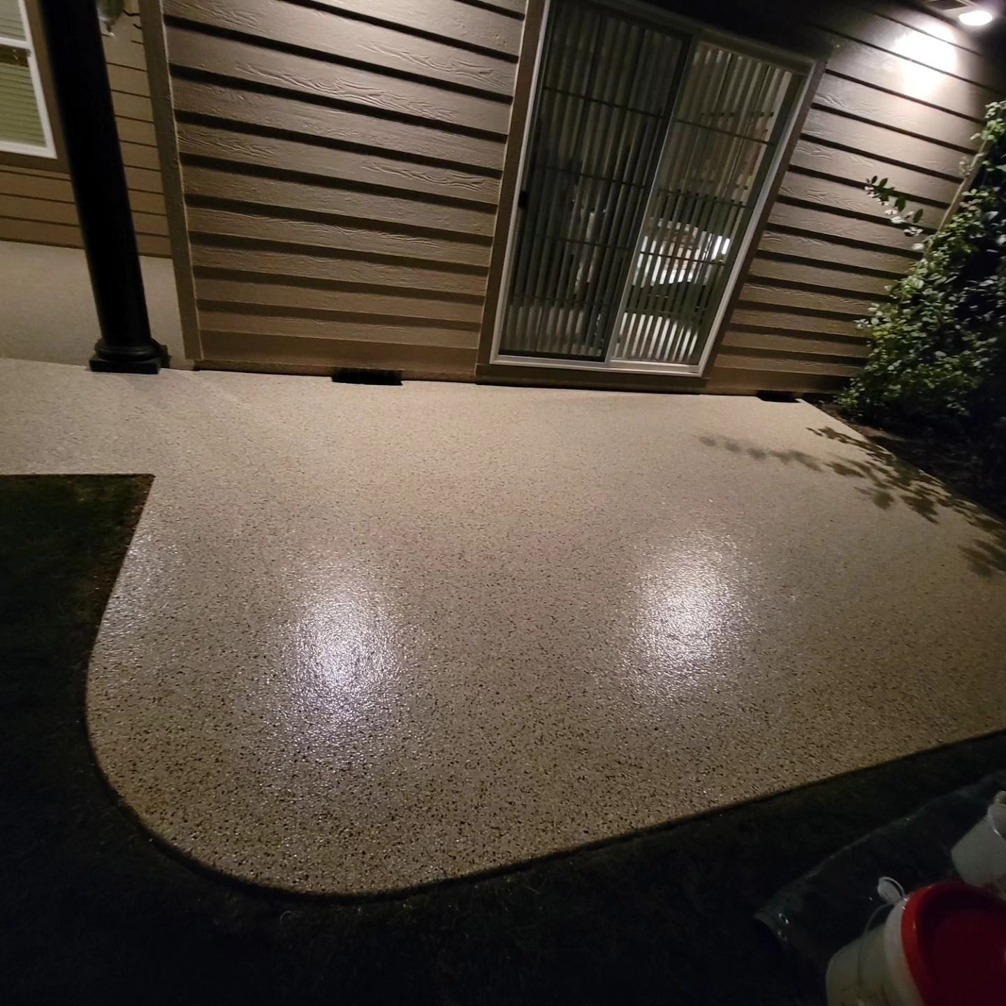 Outdoor patio at night with a speckled concrete surface, house wall, sliding door, and a potted plant.