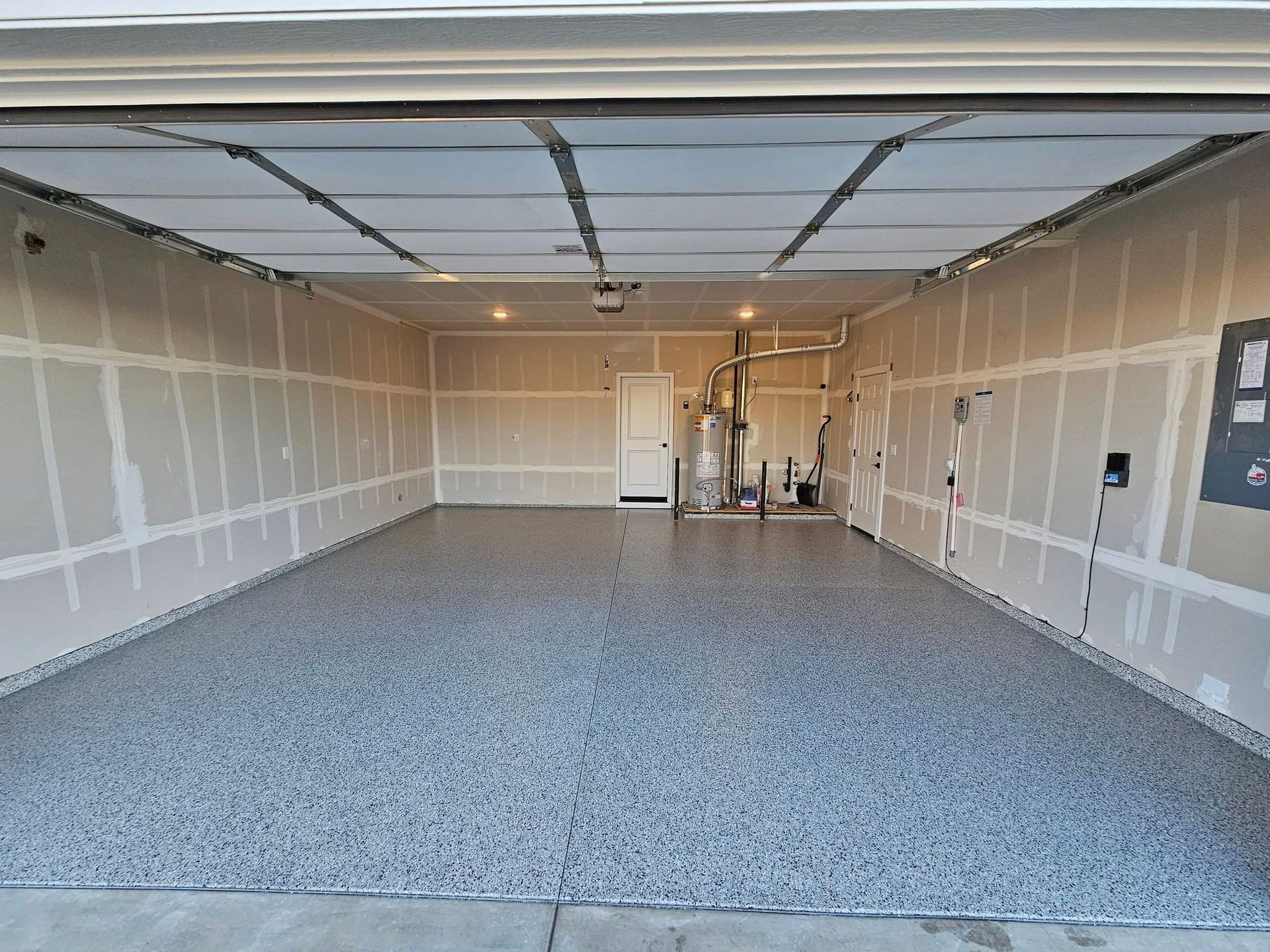 Empty garage with unfinished drywall, blue-gray speckled floor, and garage door open to the outside.