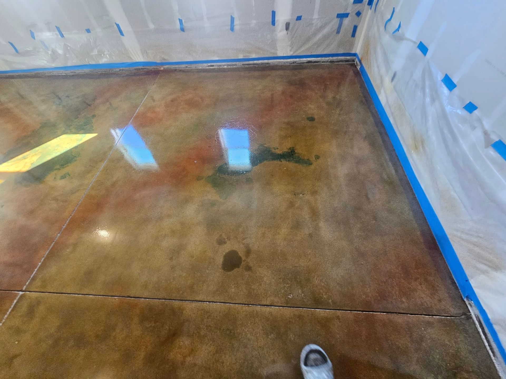 Partially finished concrete floor in a room, with blue painter’s tape along the walls and a visible shoe tip at bottom right