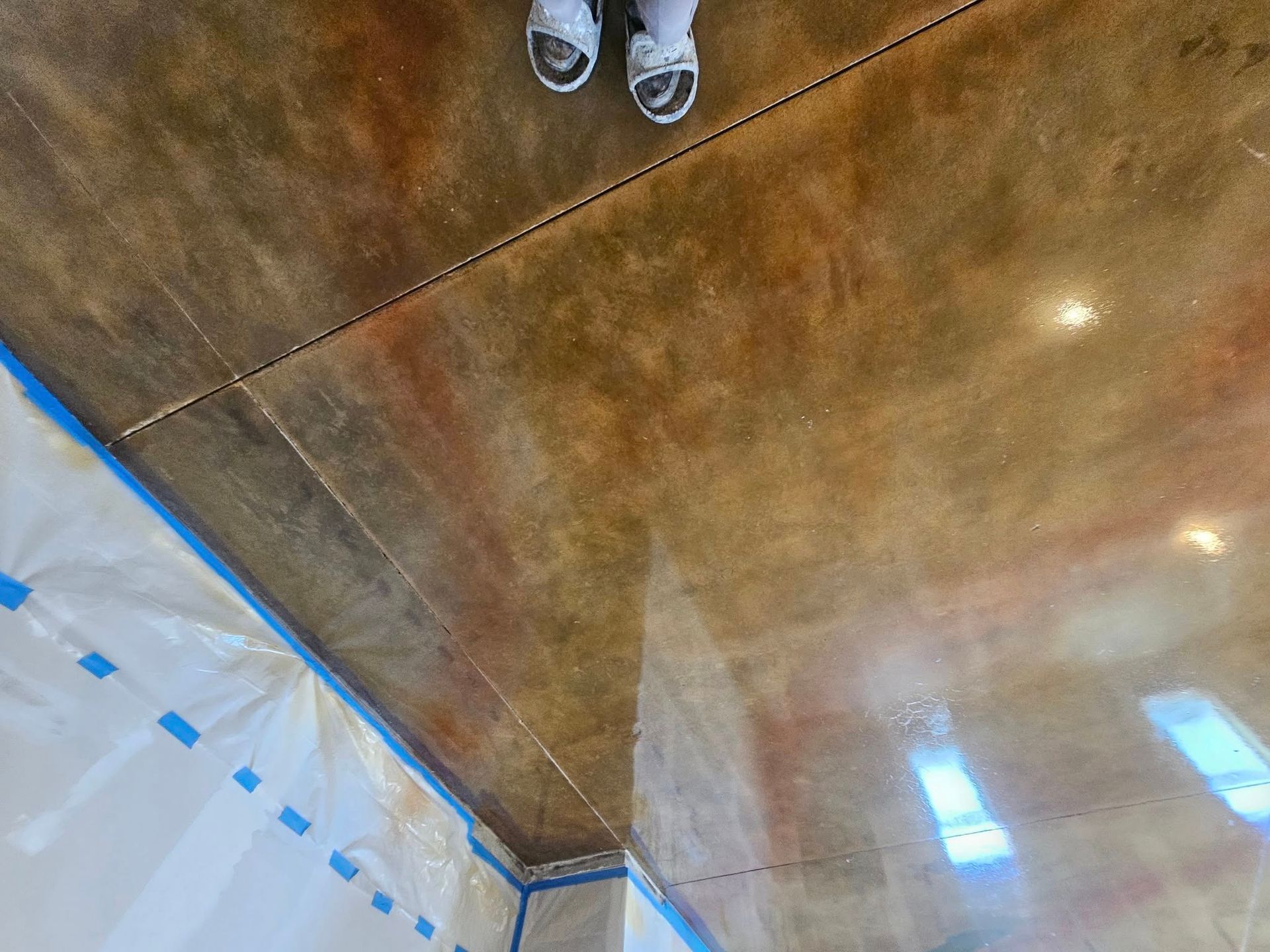 Feet standing on a brown, polished floor beside a white wall with blue tape and window reflections
