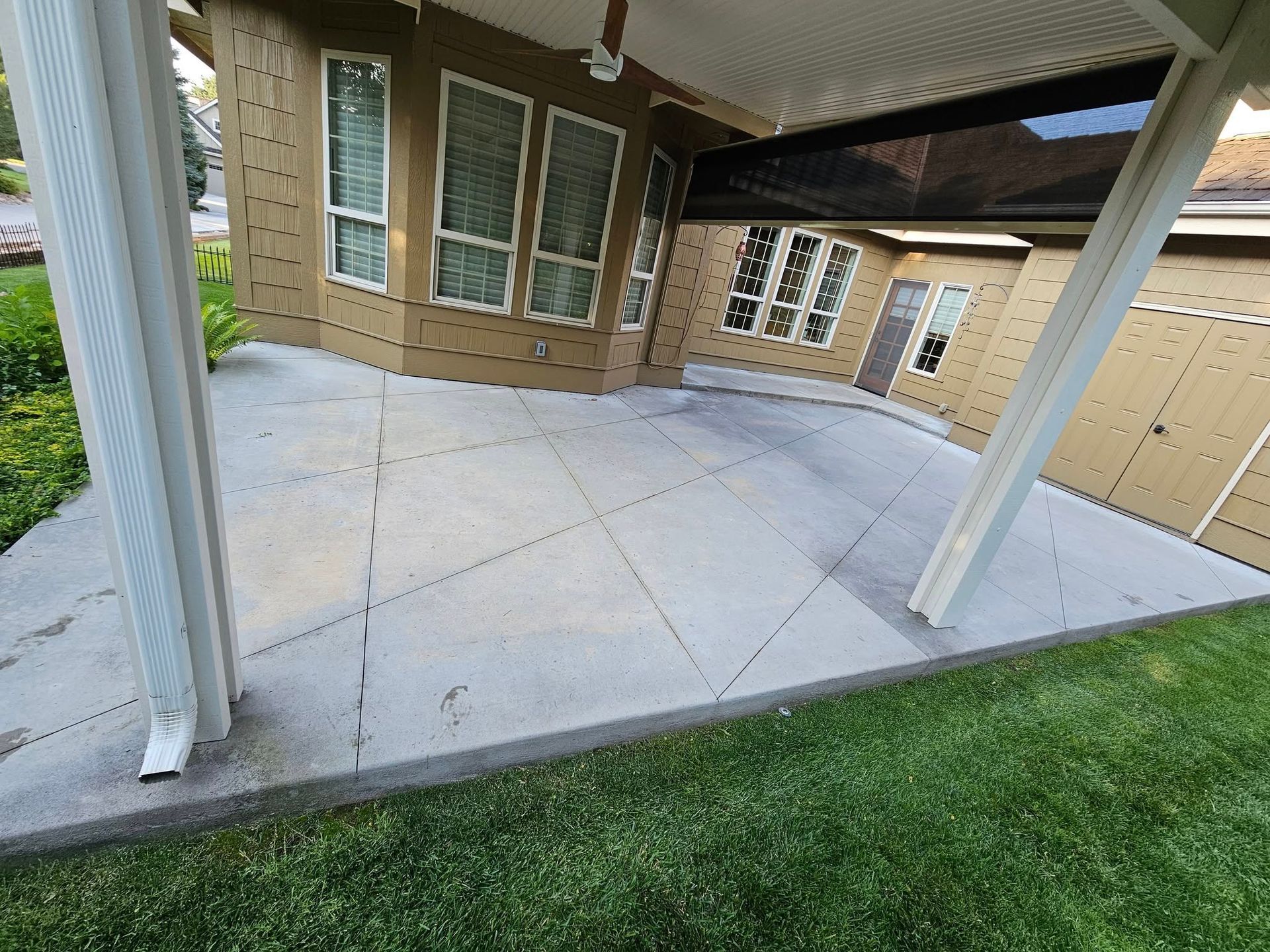 Covered concrete patio with white posts, tan siding, and large windows beside a green lawn.