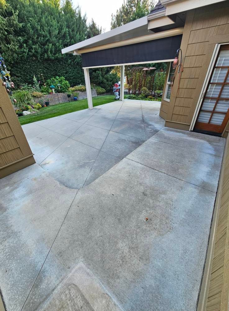Covered driveway beside a tan brick house, with a concrete patio and garden in the background.
