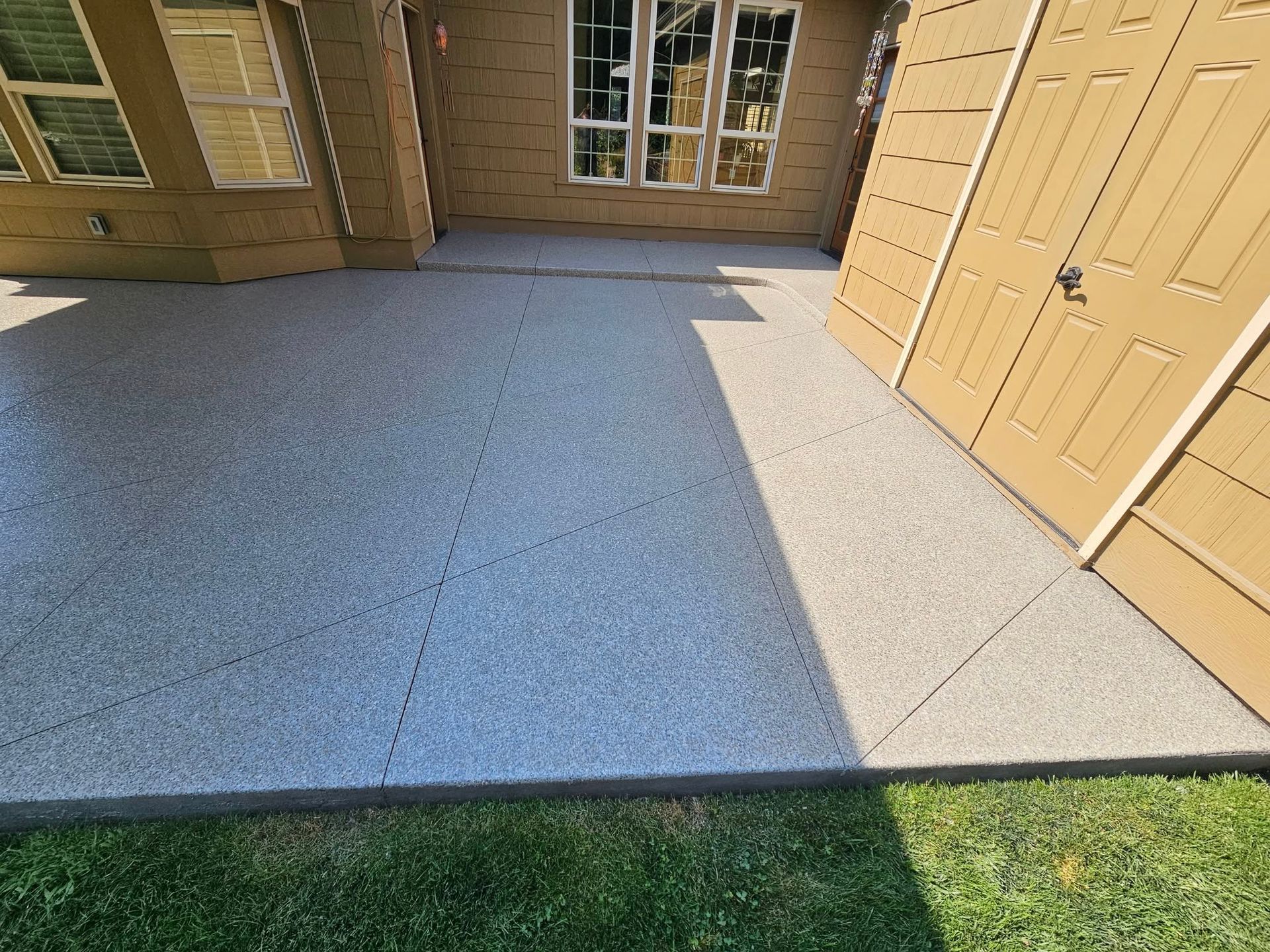 Stone patio with a strip of grass beside tan apartment doors and windows, split by sunlight and shadow