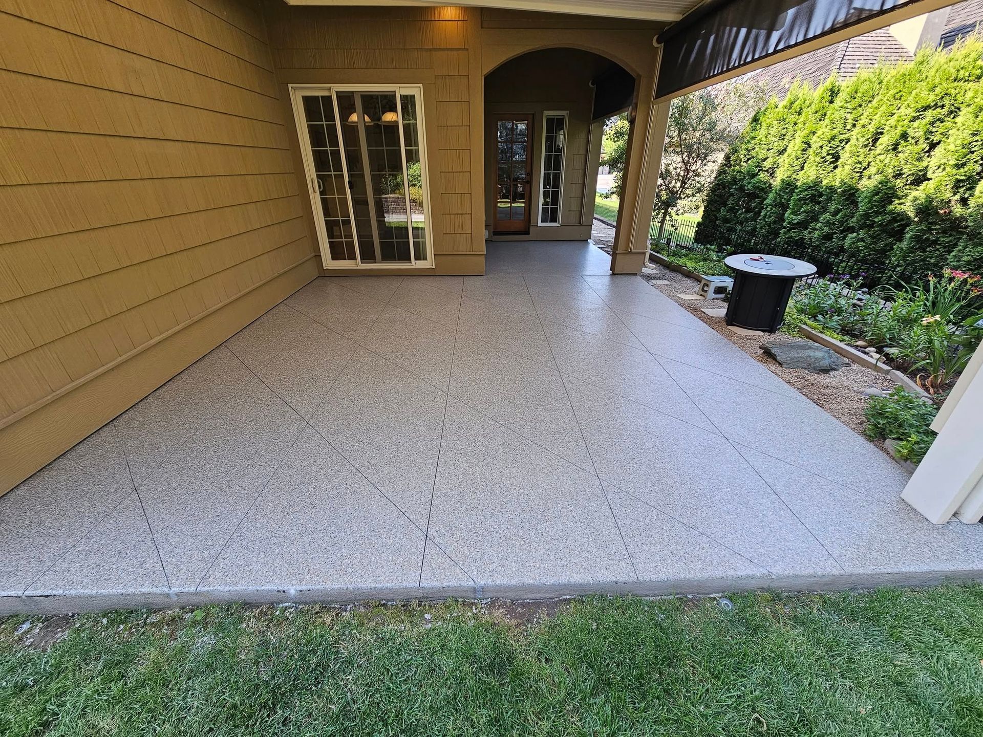 Covered patio with beige siding, speckled concrete floor, sliding glass doors, and landscaped side yard.