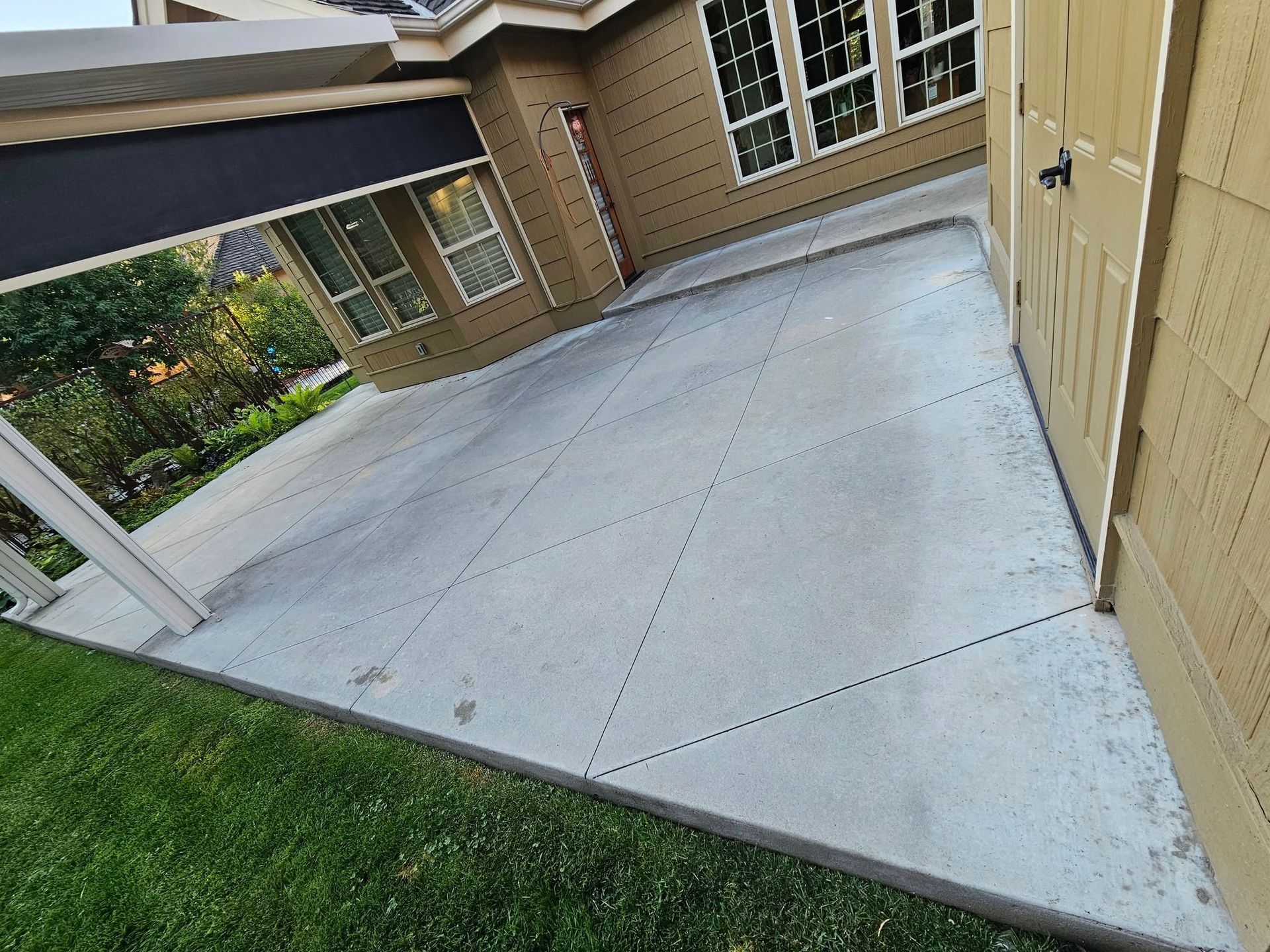 Empty concrete patio beside a beige house, with a lawn and windowed wall nearby.