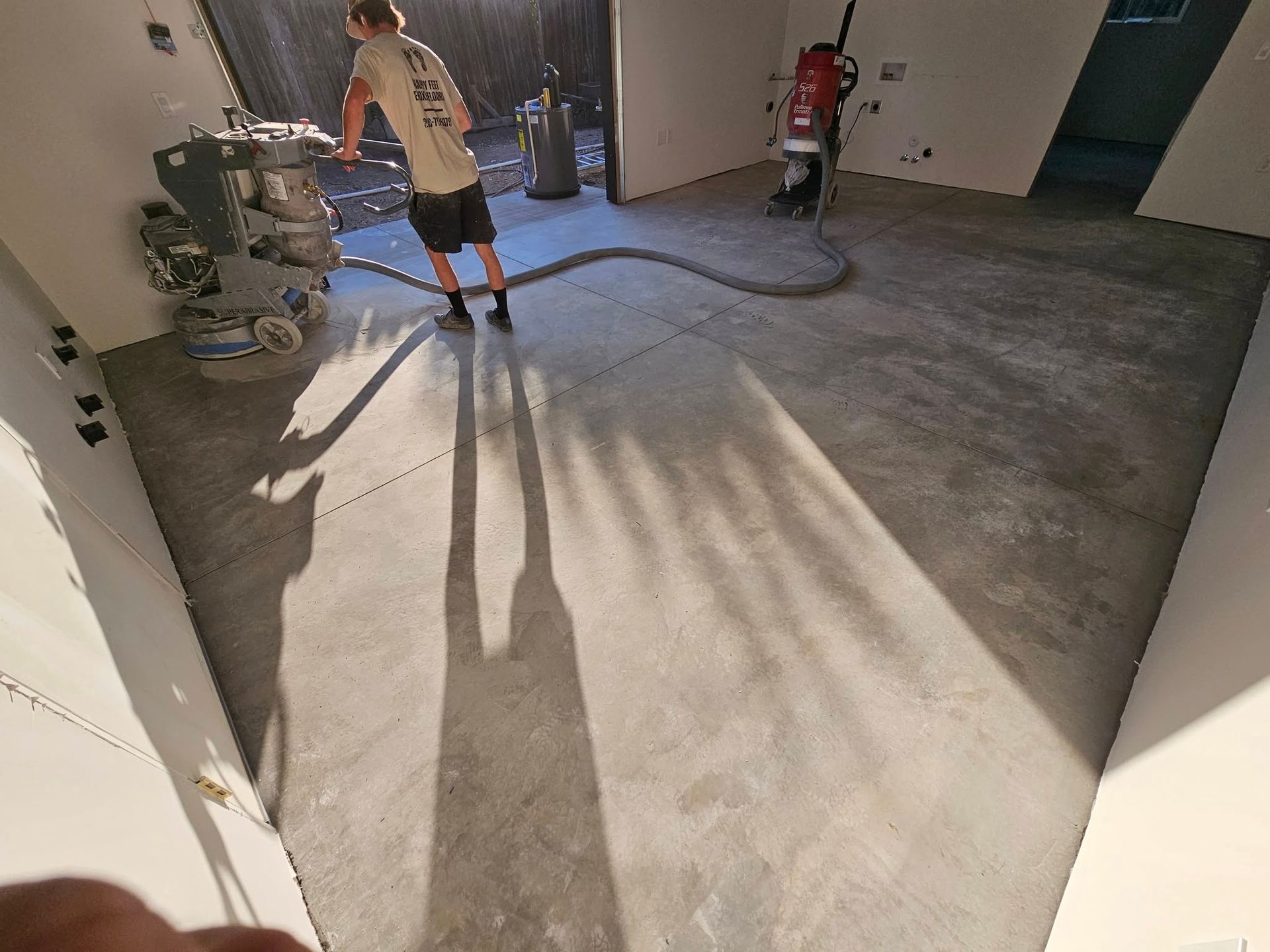 Worker using floor grinder in a bare concrete room with strong sunlight and long shadows