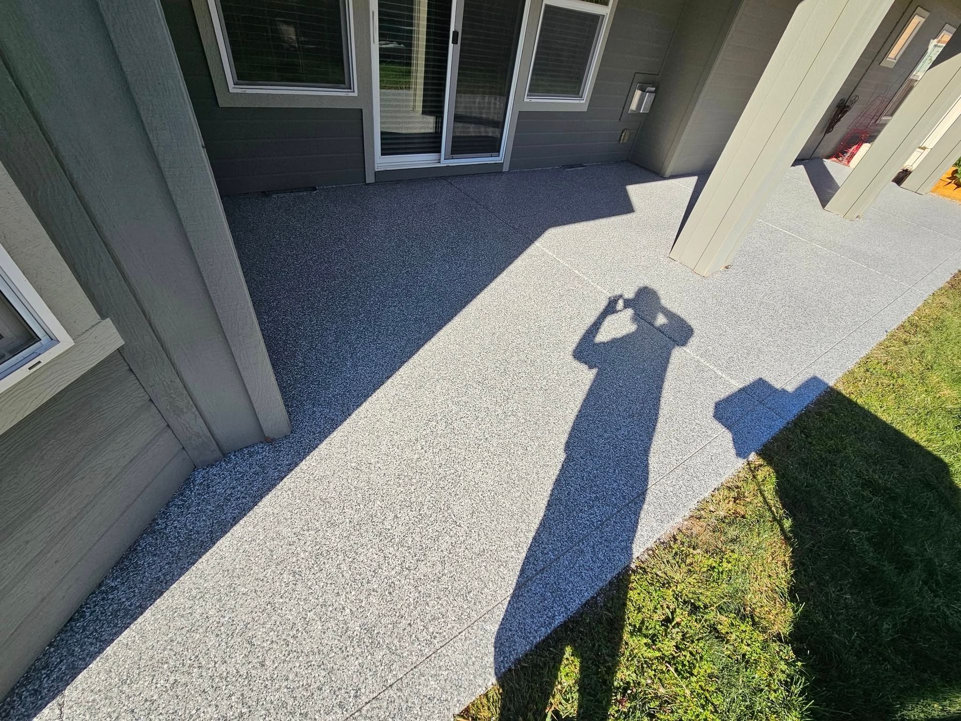 Outdoor patio with gray concrete, sliding glass doors, and a person’s shadow on the ground beside green grass.