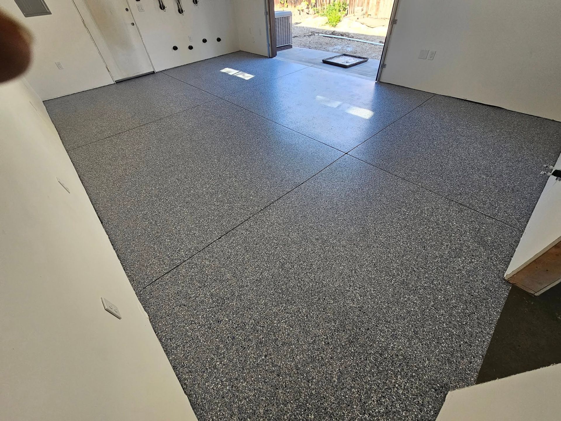 Empty room with speckled gray flooring and white walls, lit by daylight from an open doorway.