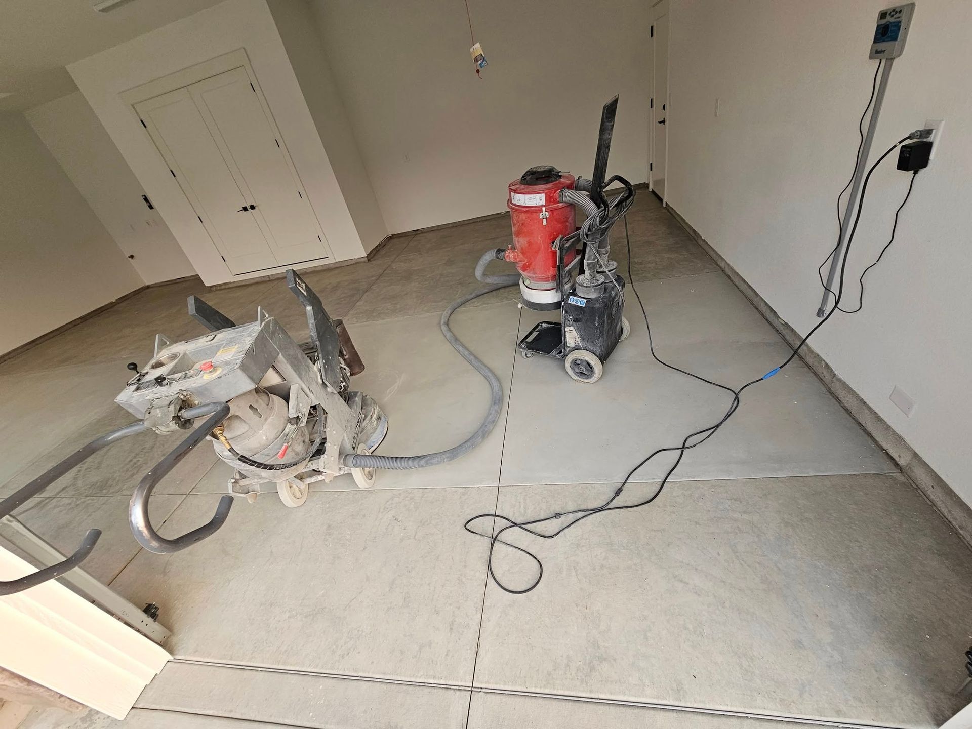 Renovation room with floor grinder, red vacuum, and cords on a bare concrete floor
