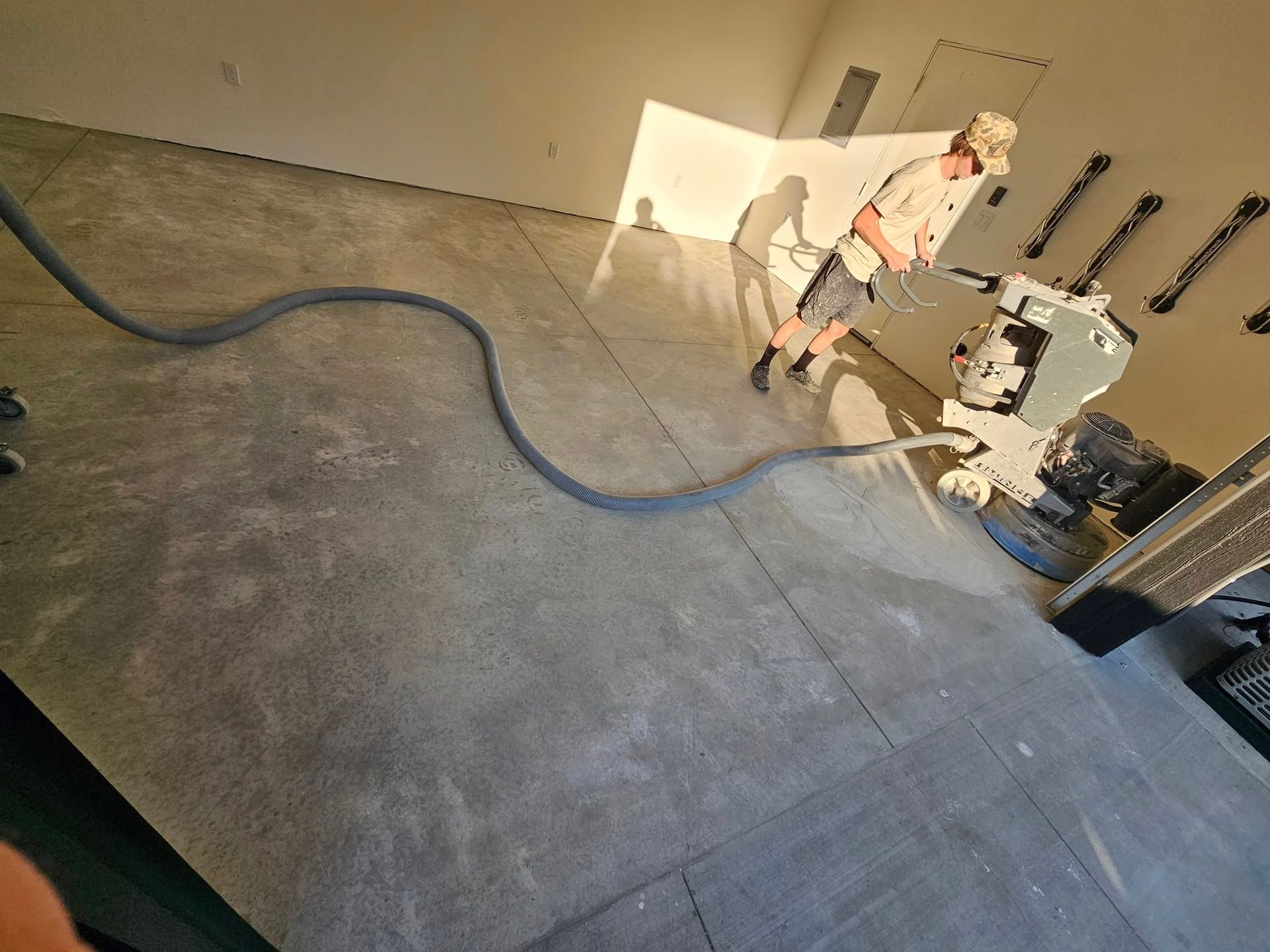Worker smoothing concrete floor with a machine in a bare room, hose stretched across the floor.