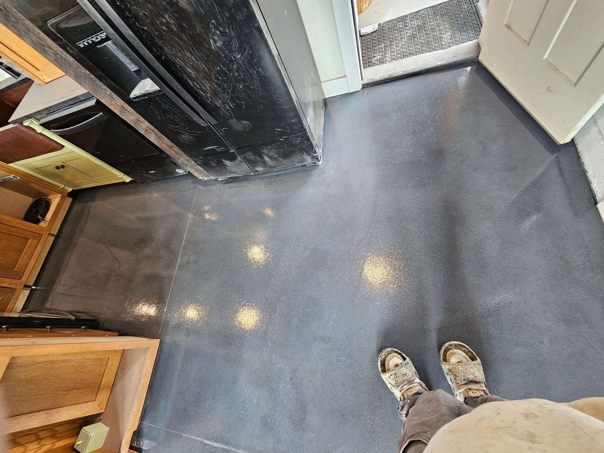 Top-down view of a gray kitchen floor with cabinets, doorway, and a person’s shoes at the bottom edge