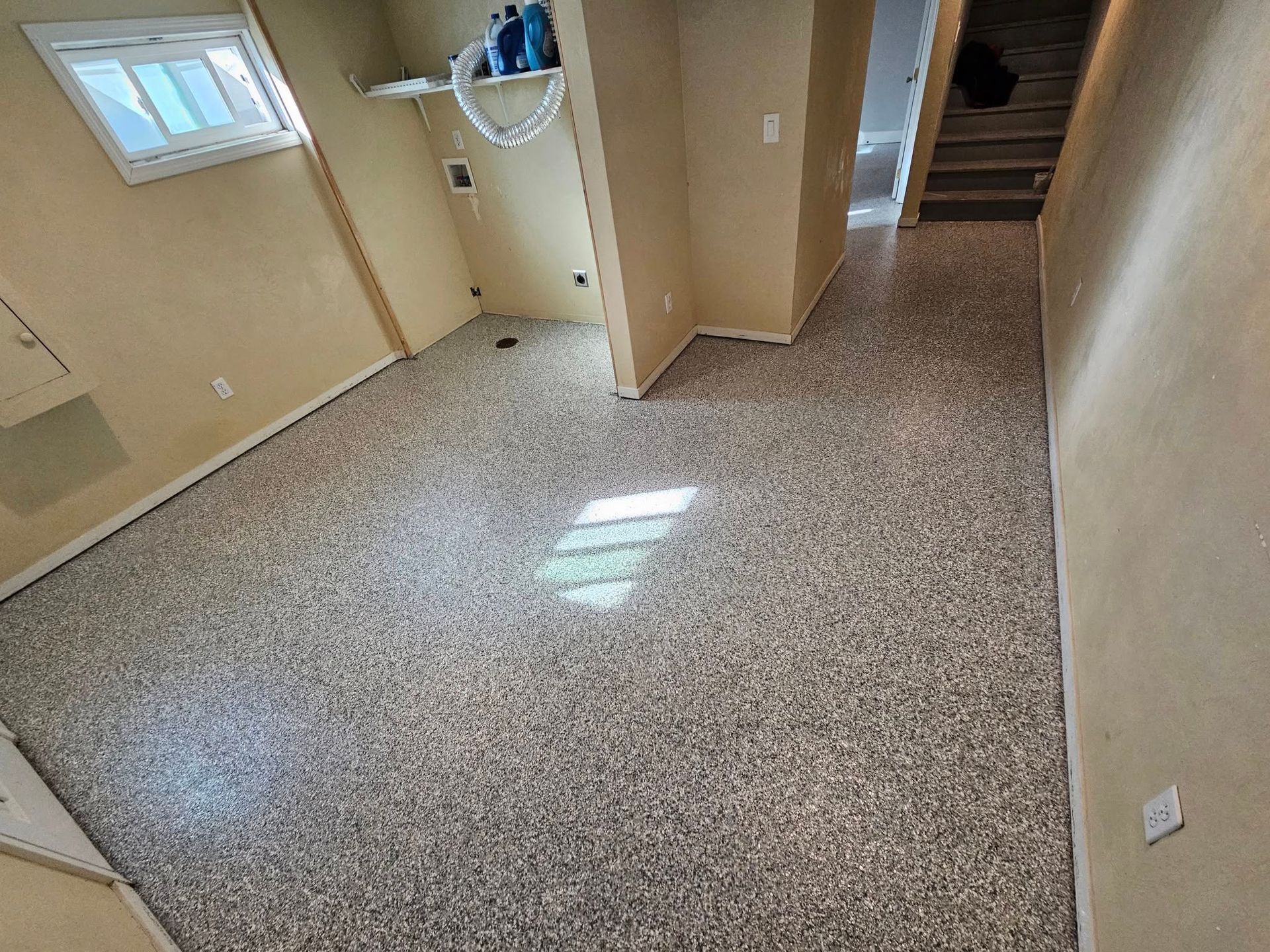 Empty beige basement room with speckled carpet, small window, and stairs leading up.
