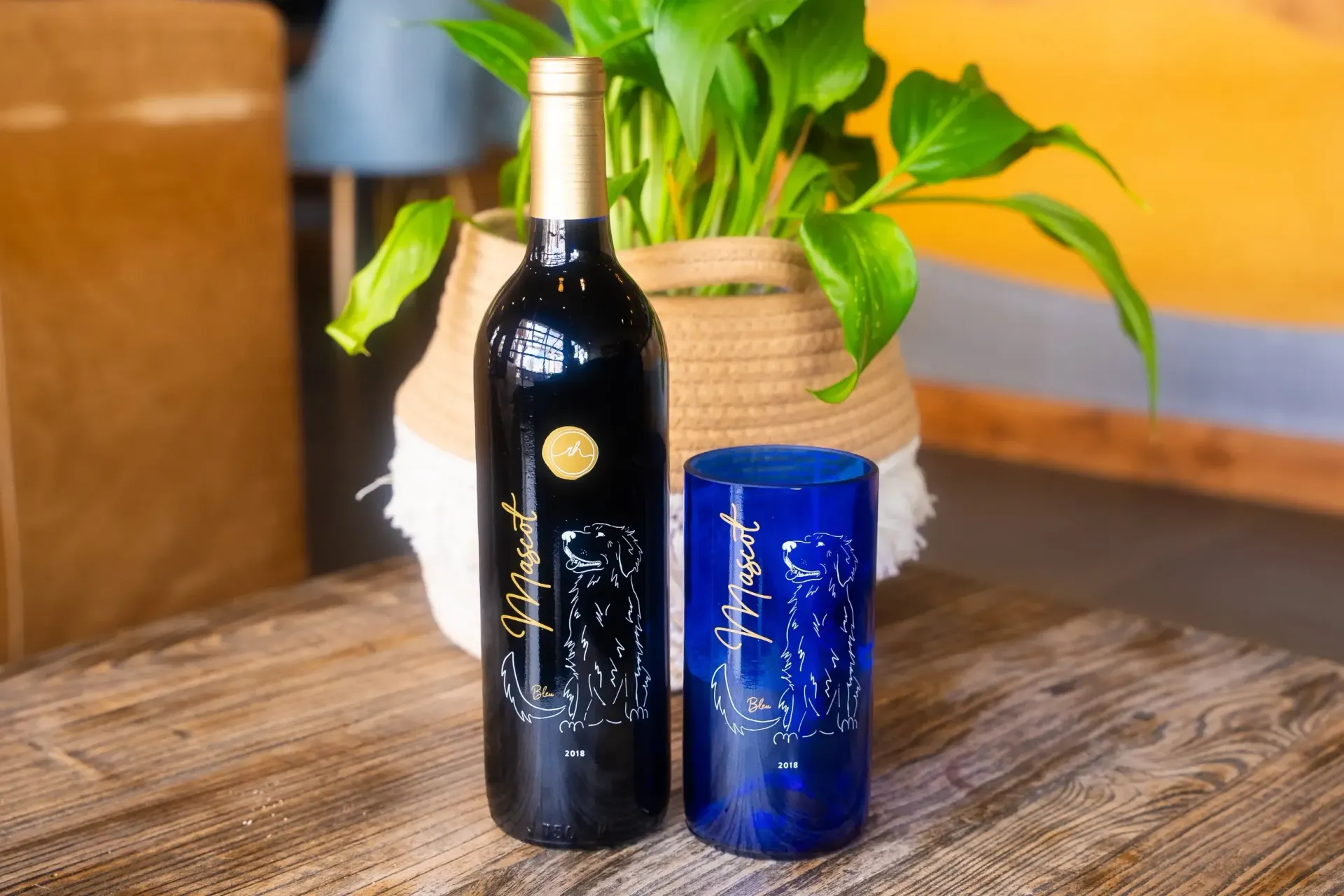 Wine bottle and blue glass on a wooden table, with a potted plant in the background.