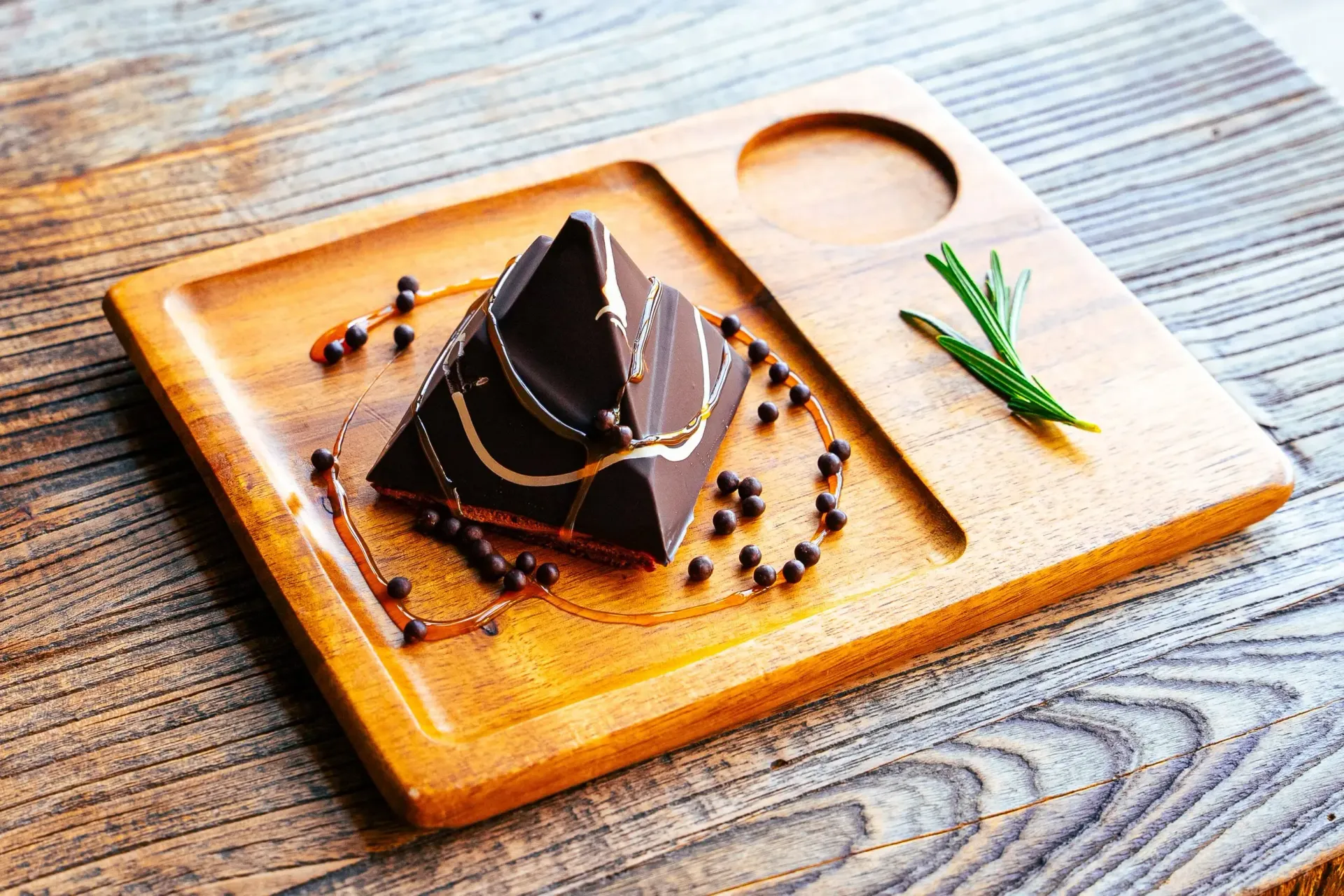 Dark chocolate pyramid dessert on a wooden tray, garnished with chocolate beads and a sprig of rosemary.
