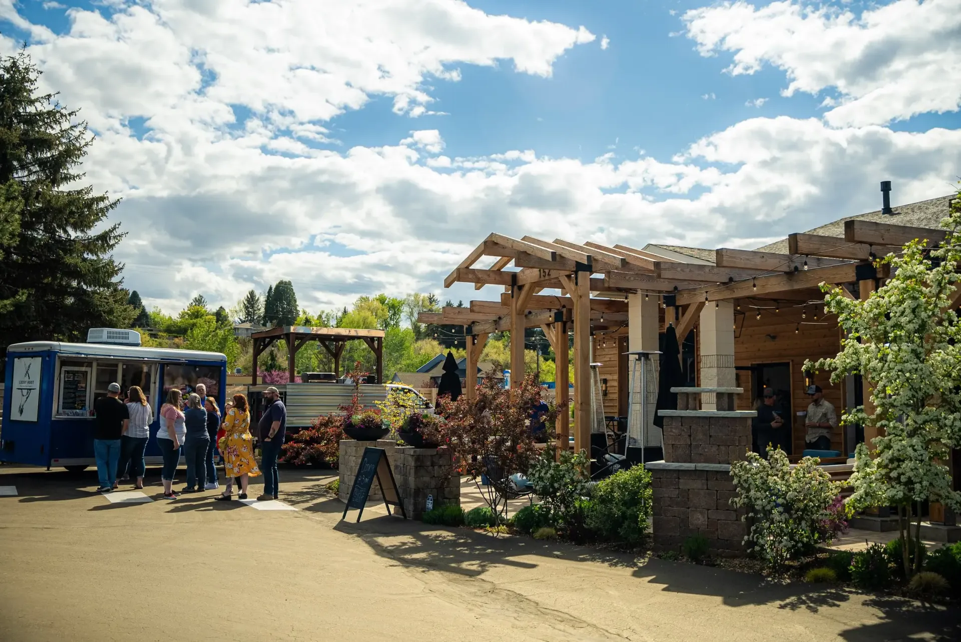 People outside a building with a pergola, next to a blue food truck on a sunny day.
