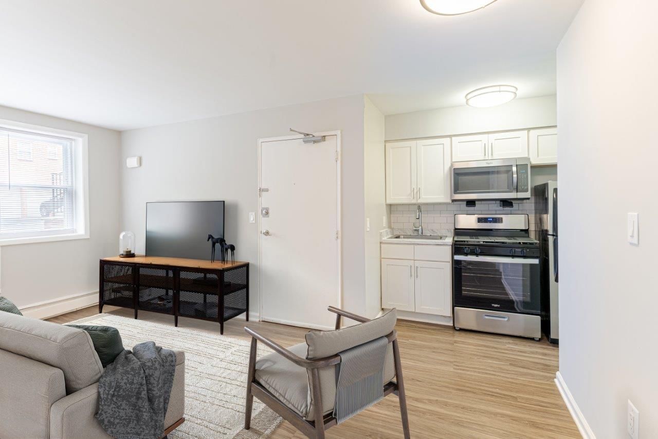 Bright living room with gray sofa, TV stand, and open kitchen with white cabinets and stainless steel appliances