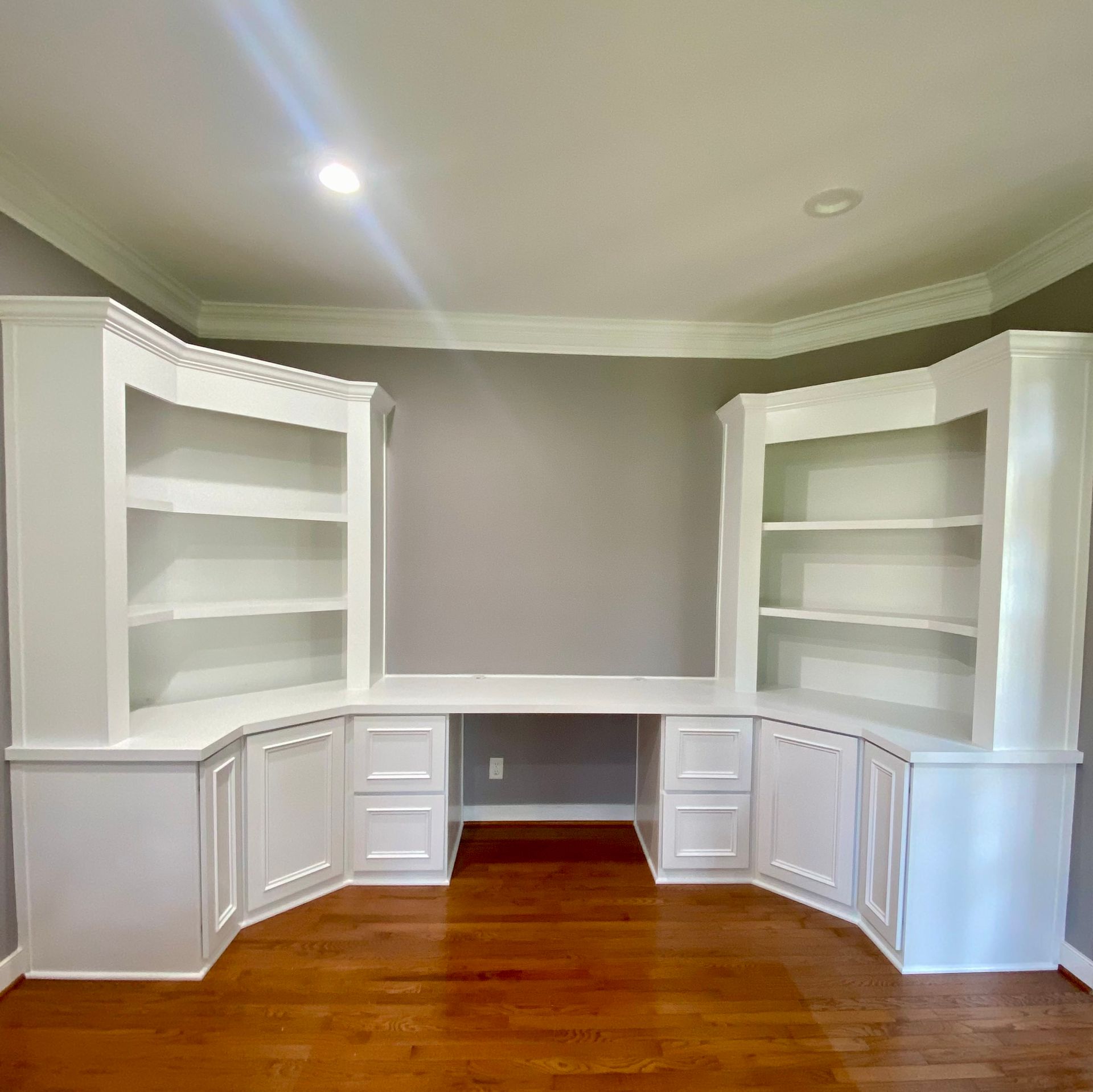 Built-in white corner shelving and cabinets in a gray room with hardwood floors, under a tray ceiling.