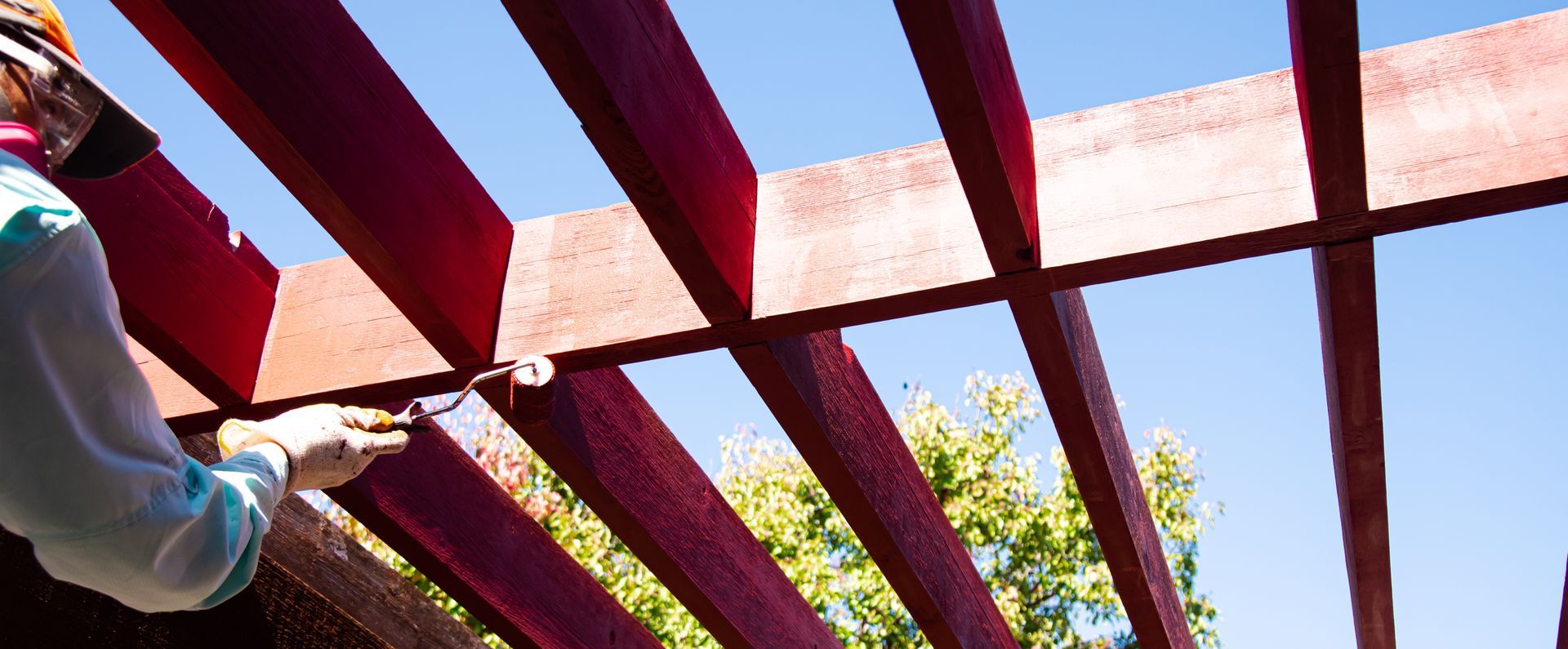 Person painting red beams of a pergola against a bright blue sky.