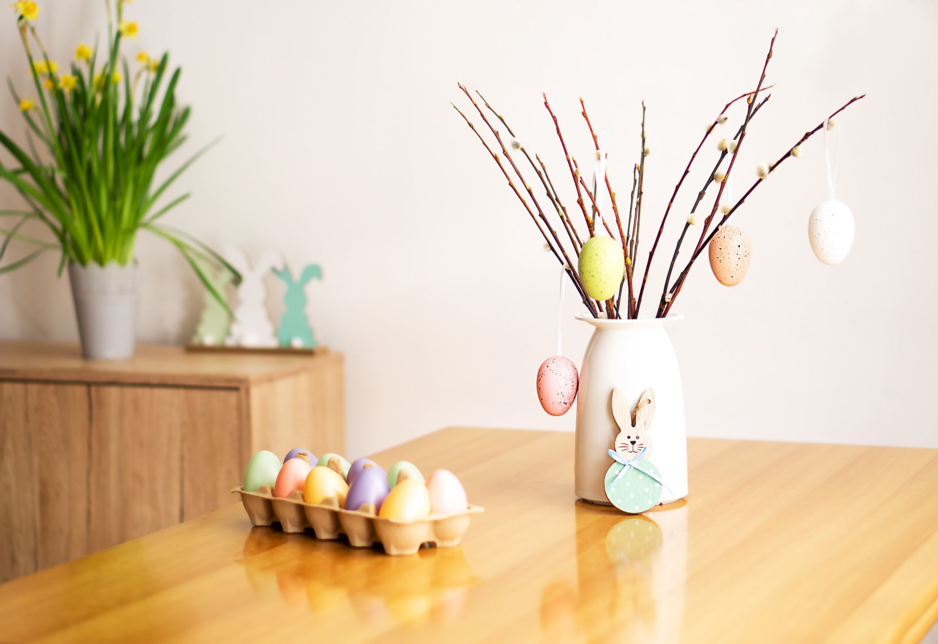Easter table decor: painted eggs in a carton, vase with pussy willow branches, and decorative bunny.