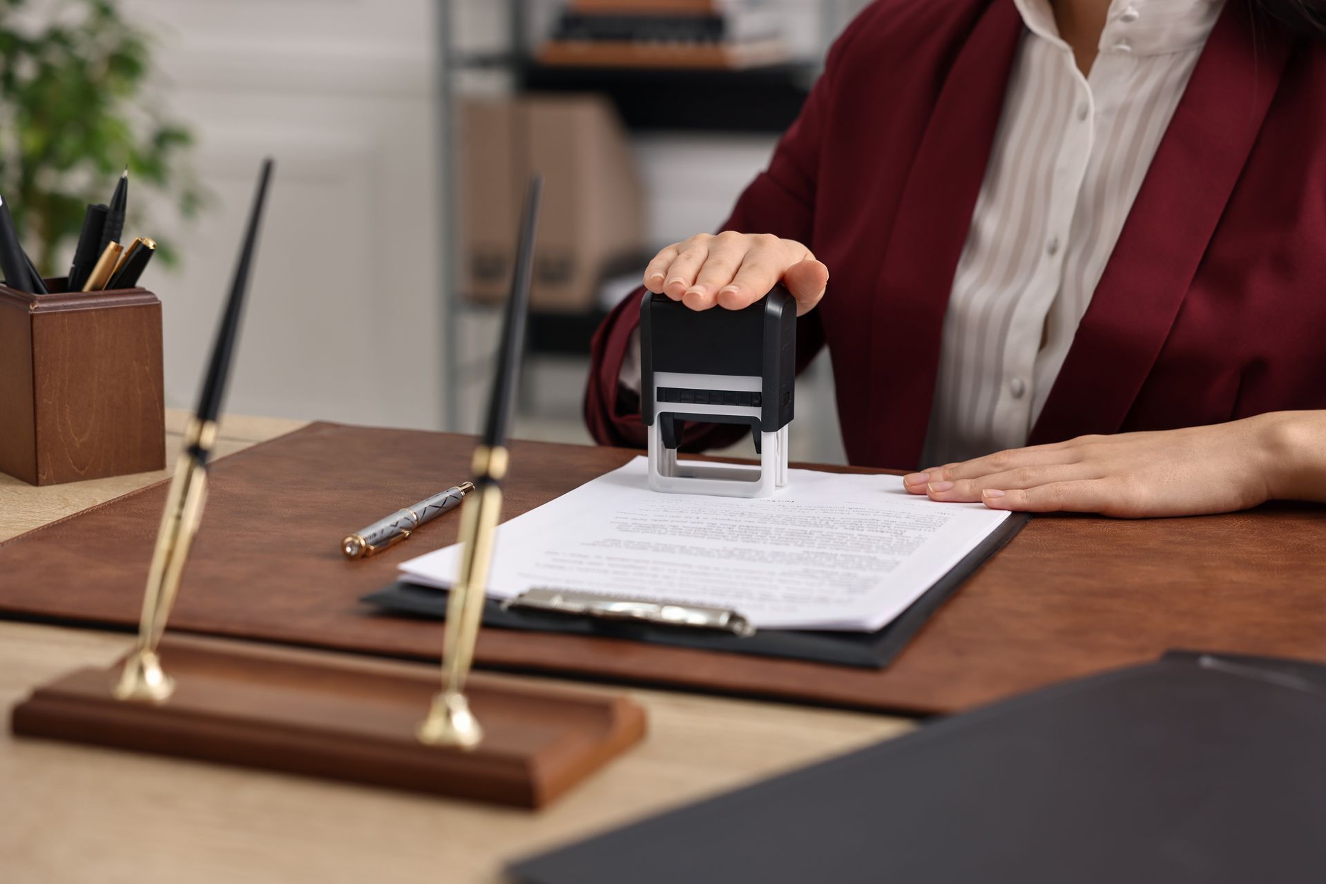 Woman in maroon blazer stamps a document on a desk with pen holders, pens, and a black file.