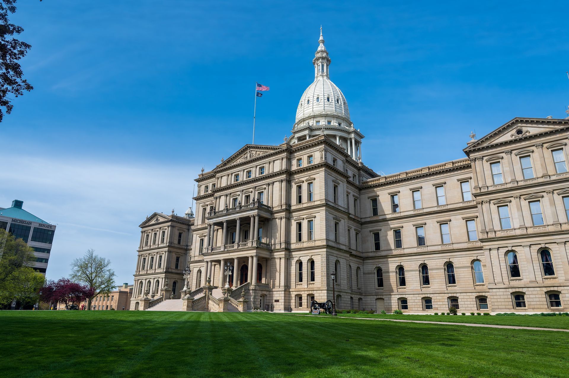 Historic capitol building with a white dome and flag, seen from a green lawn under a blue sky