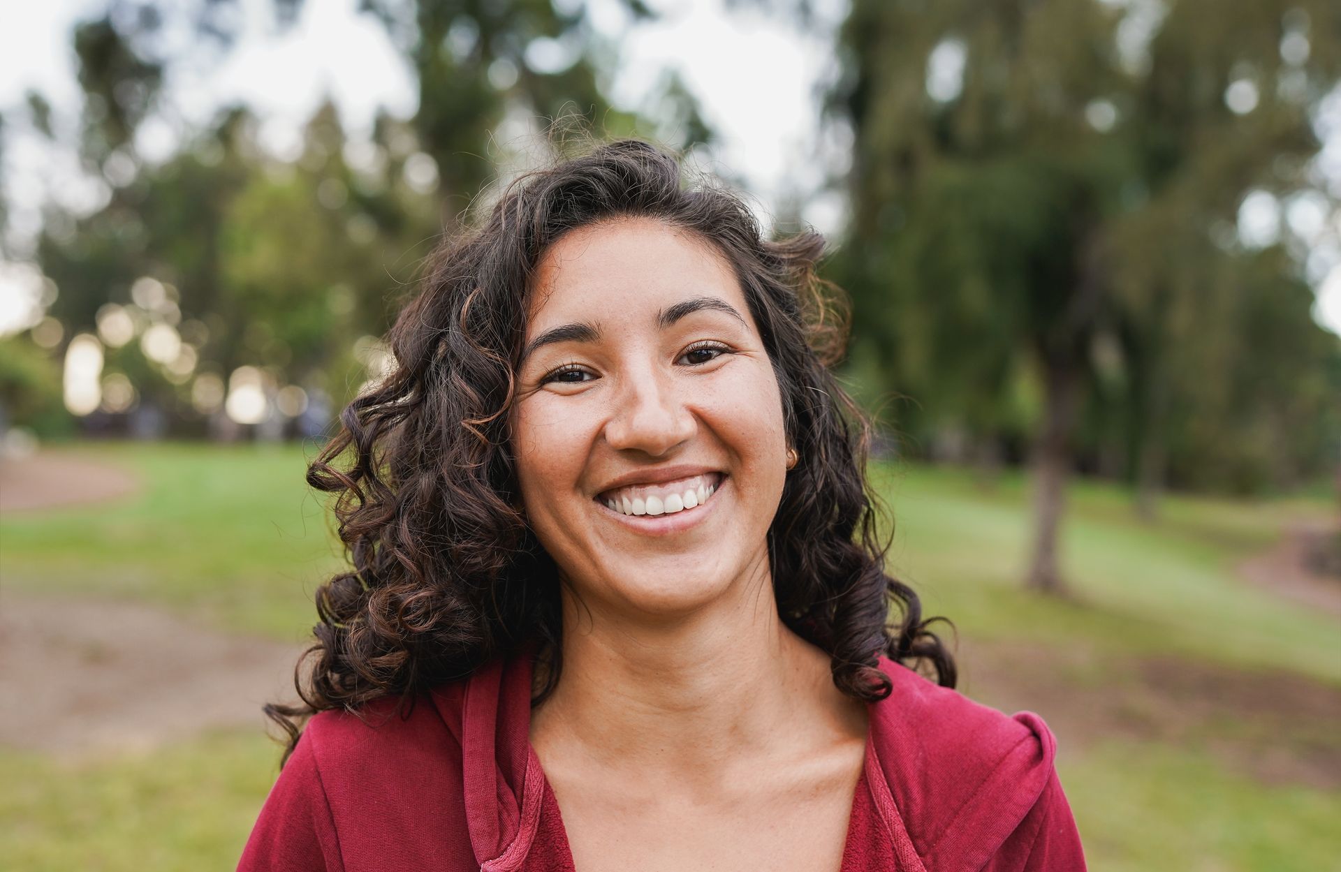 A young Native American woman with curly hair is smiling in a park.